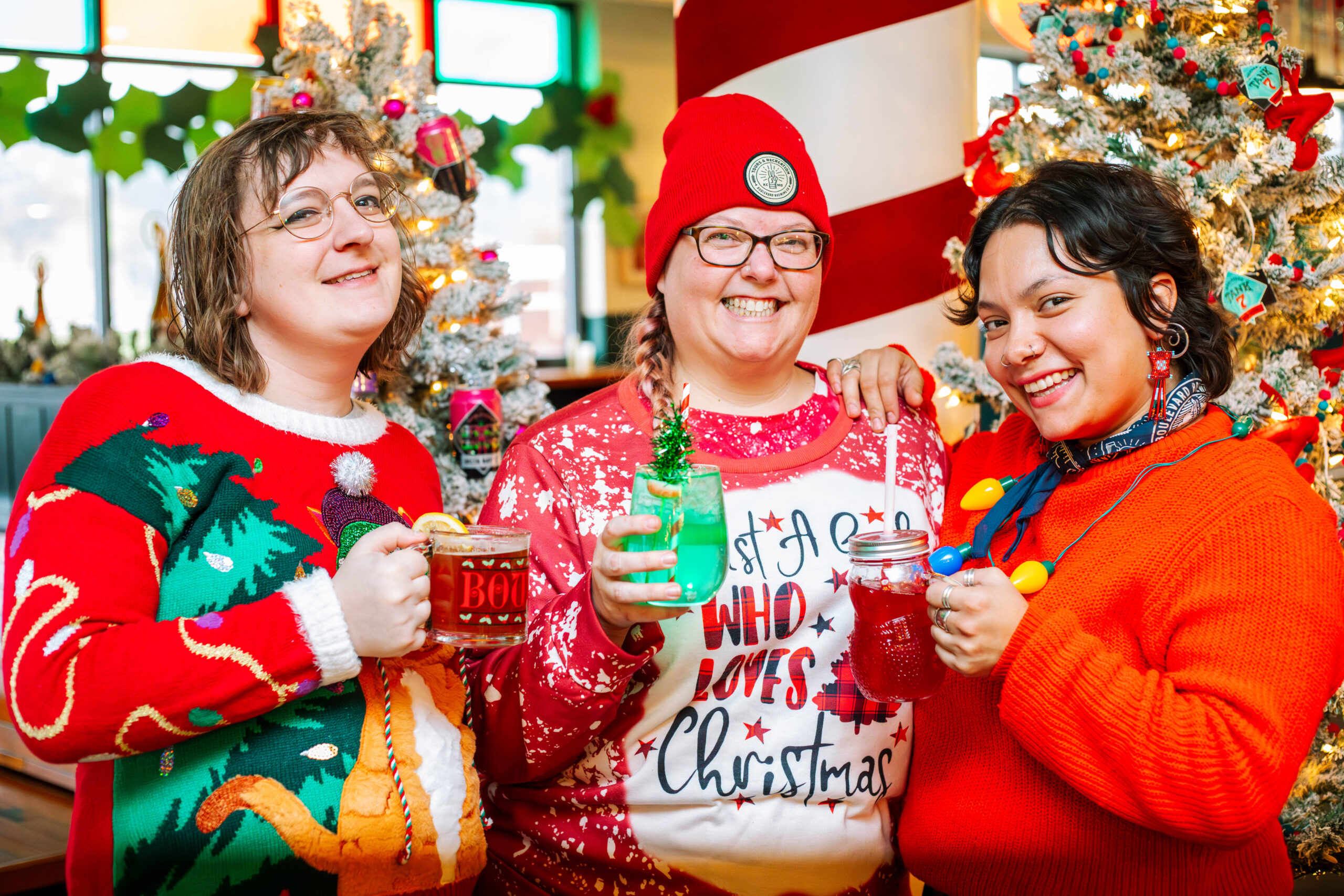 Three people in colorful Christmas sweaters smile and hold festive holiday drinks. Decorated Christmas trees and holiday ornaments are in the background, creating a cheerful, festive atmosphere.