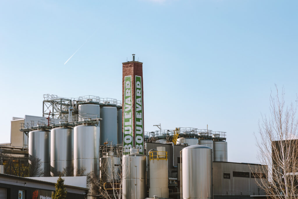 A brewery with multiple large silver fermentation tanks and a tall red chimney displaying a vertical green "Boulevard" sign under a clear blue sky, perfect for enjoying football season.