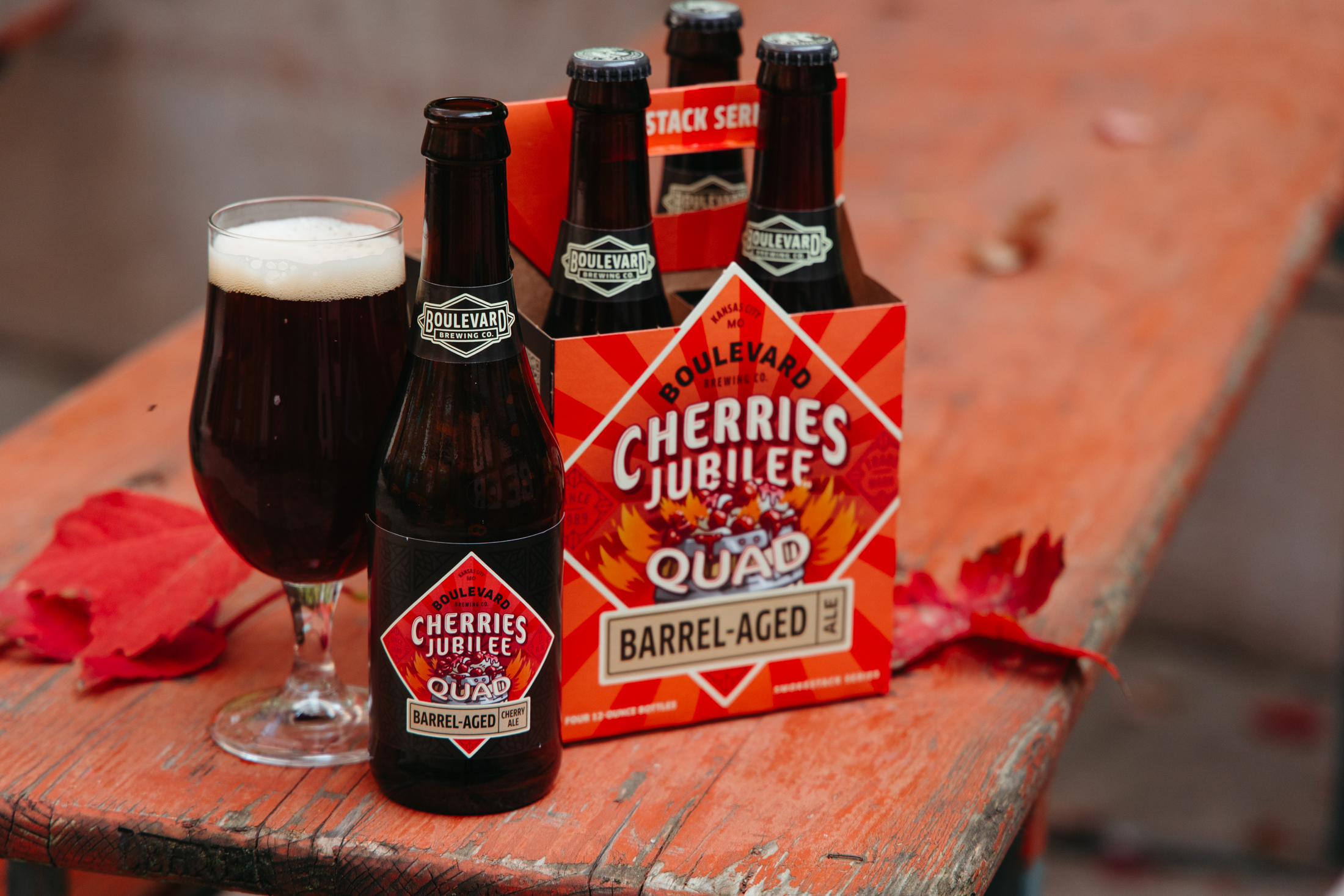 A glass of dark beer sits beside a bottle and a four-pack of Boulevard’s Cherries Jubilee Quad Barrel-Aged beer on a wooden table with scattered red autumn leaves.