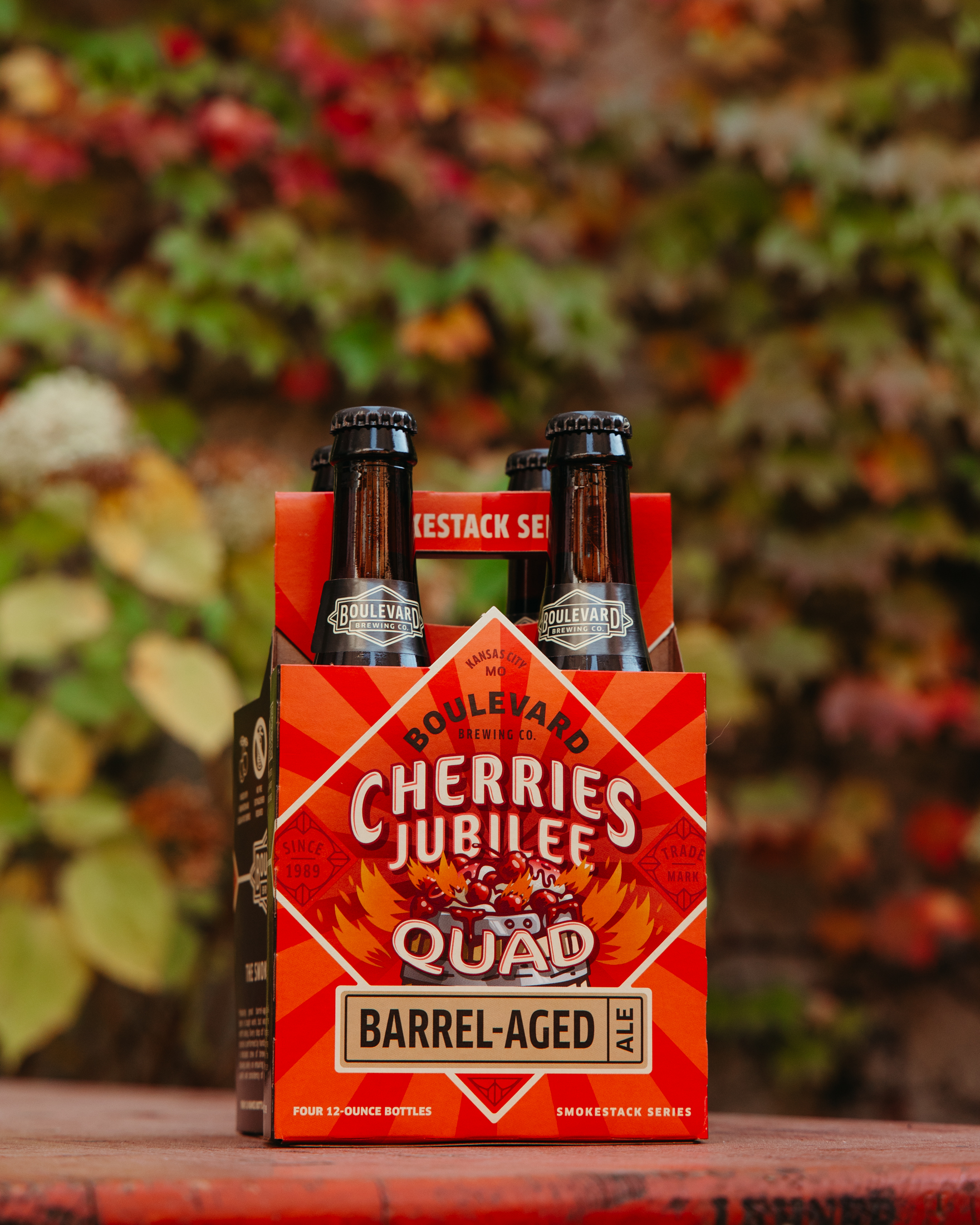 Four bottles of Boulevard Brewing Co.’s Cherries Jubilee Quad Barrel-Aged Ale in a red and orange cardboard carrier, placed on an outdoor surface with blurred autumn foliage in the background.
