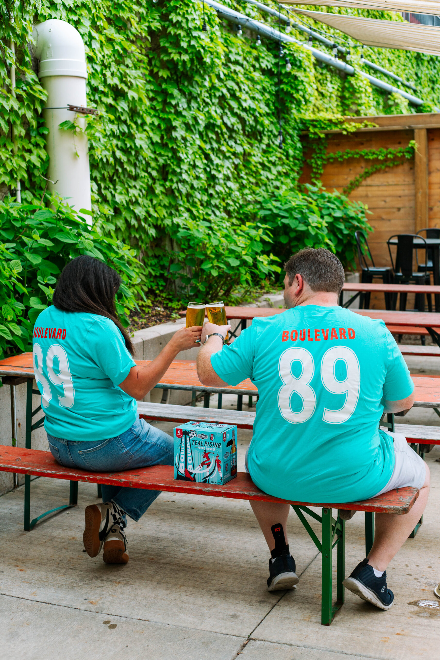 Two people in matching turquoise "Boulevard 89" shirts sit on a bench at an outdoor table, toasting with beer glasses. Green vines climb the walls, while a box of Boulevard beer sits on the table—perfect for celebrating after a football game.