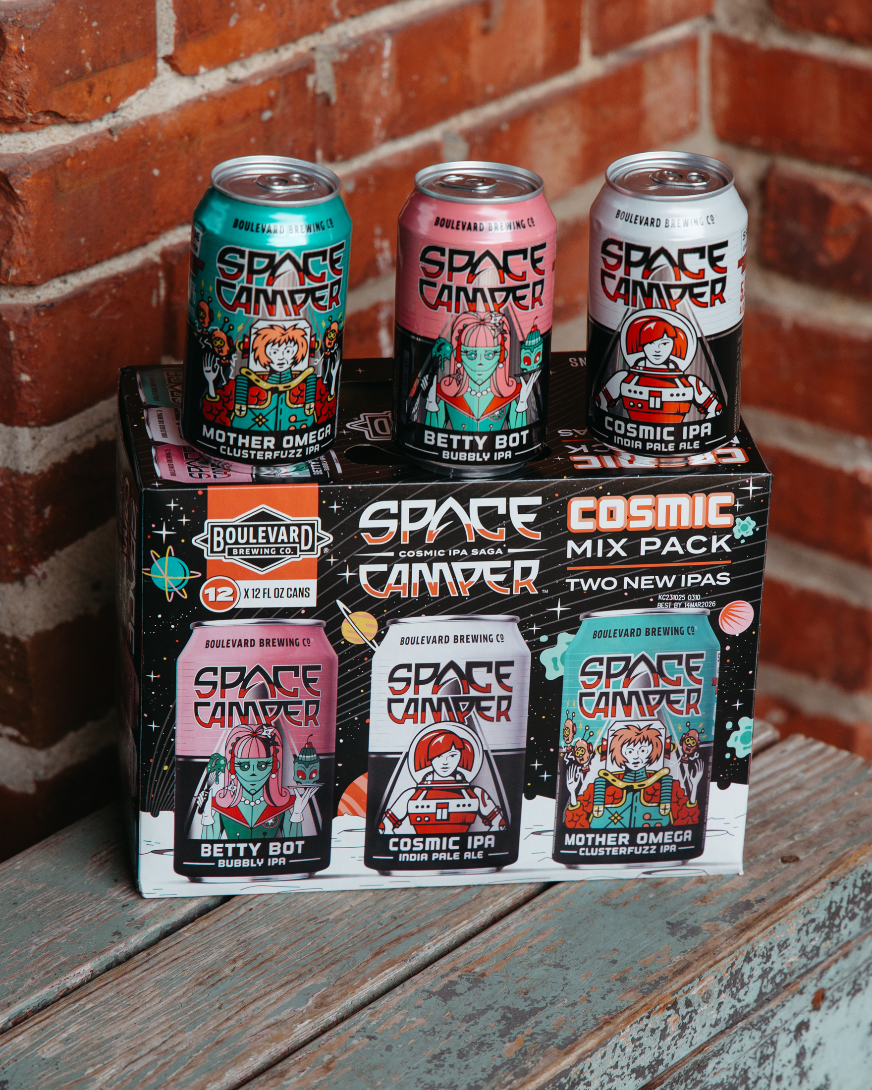 A 12-pack box of Boulevard Brewing Co.’s Space Camper Cosmic Mix Pack, featuring colorful cans of “Betty Bot” and “Mother Omega” IPAs, displayed on a wooden bench against a brick wall.