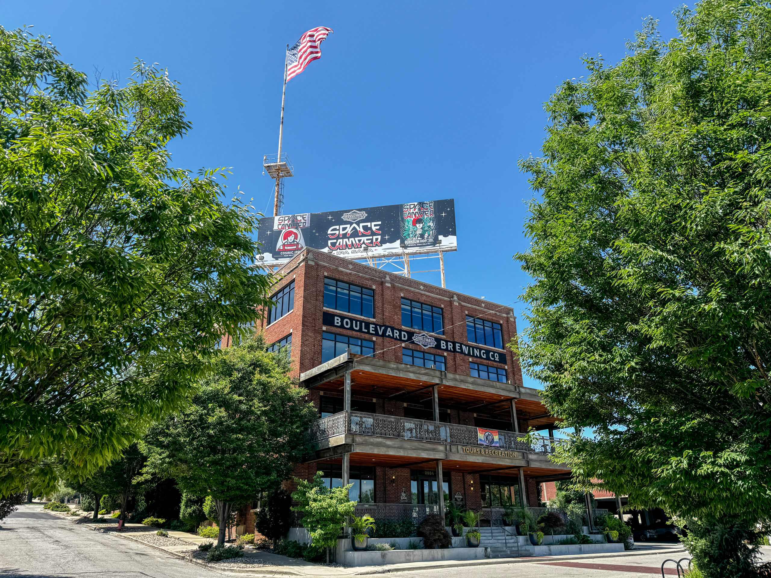 A large brick building with "Boulevard Brewing Co." on the front, surrounded by green trees, features a U.S. flag and a "Space Camper" billboard atop the roof—perfect for relaxing after a football game—under a clear blue sky.