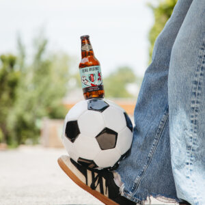 A person wearing jeans and black sneakers balances a football with a bottle of Teal Rising beer resting on top of the ball, outdoors on a sunny day.