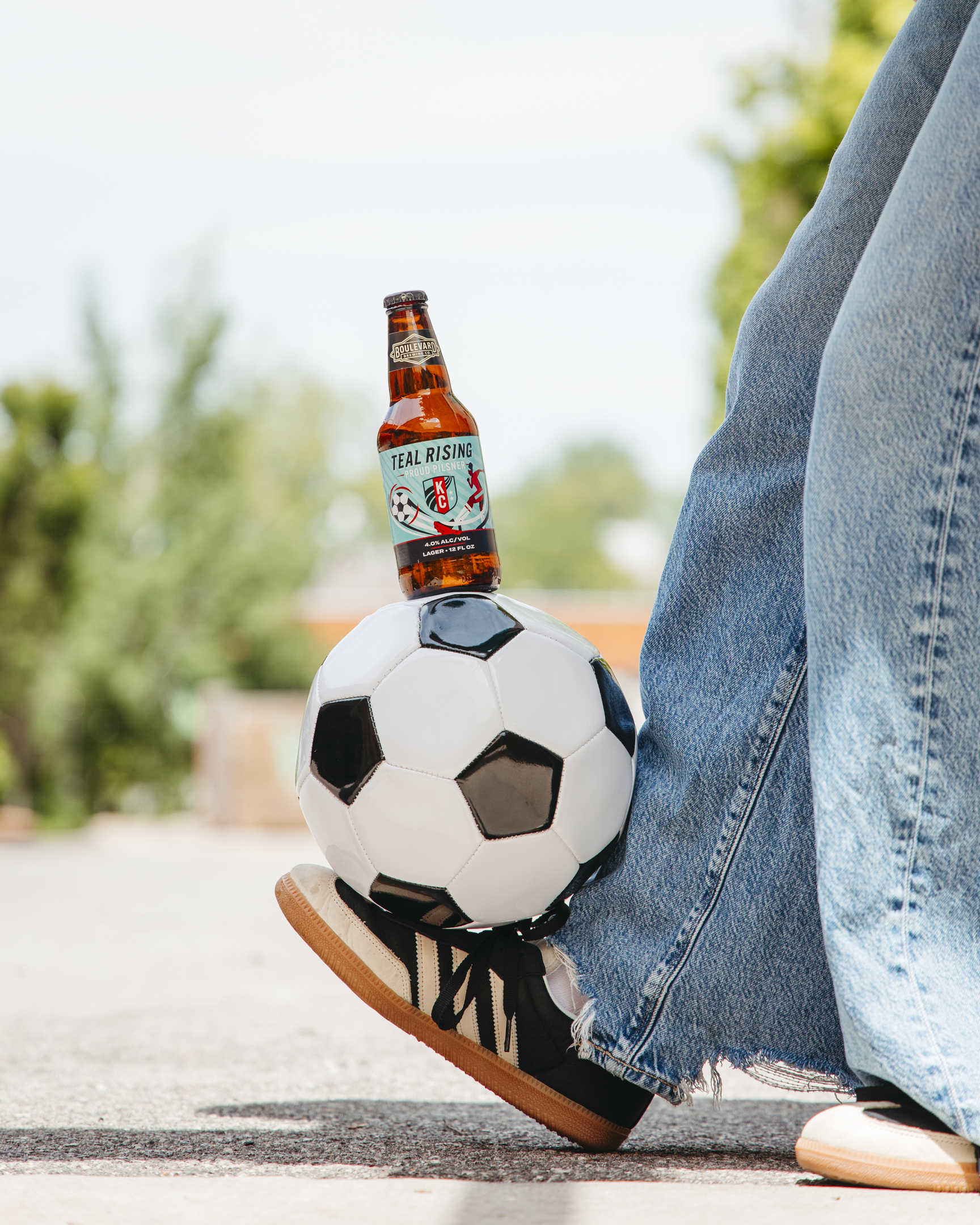 A person wearing jeans and black sneakers balances a football with a bottle of Teal Rising beer resting on top of the ball, outdoors on a sunny day.