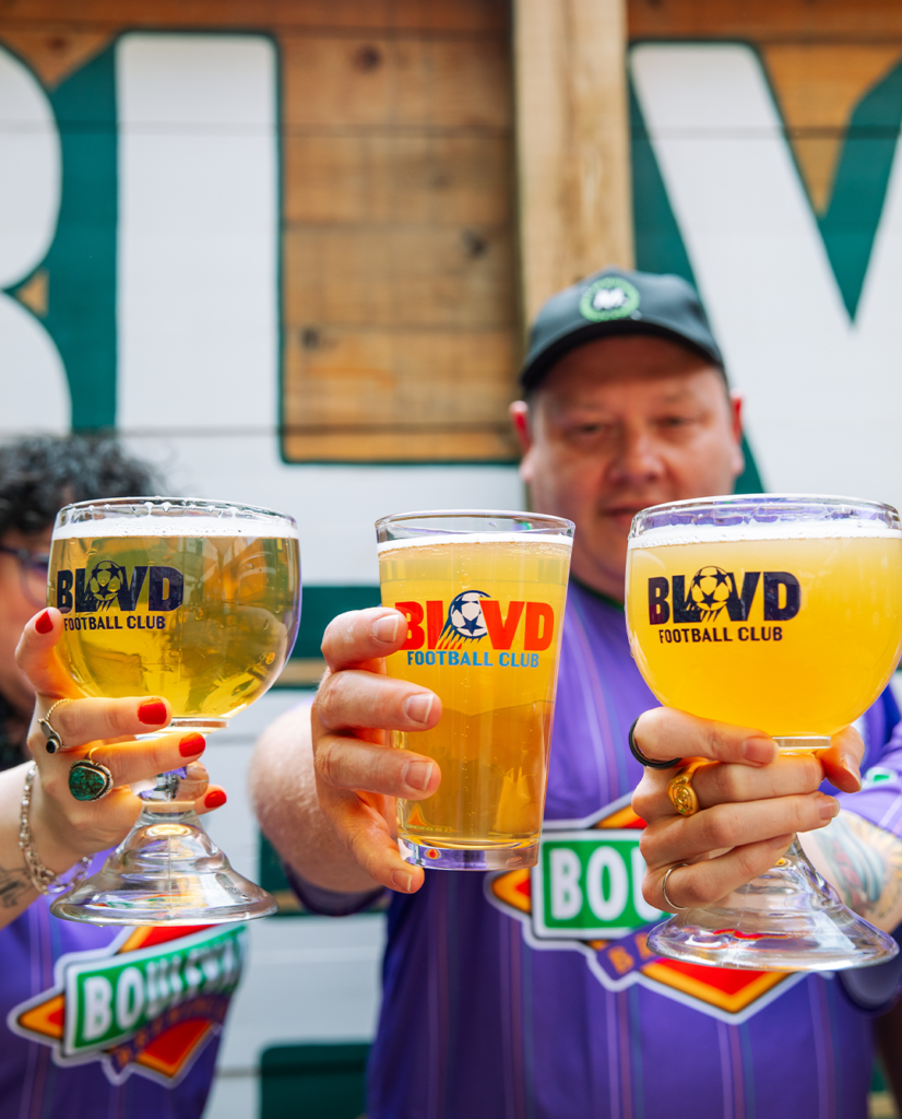 Three people in purple jerseys hold up large glasses of beer with "BLVD Football Club" logos. Gathered outdoors in front of a mural, their drinks range from light to golden yellow, celebrating their love for football together.