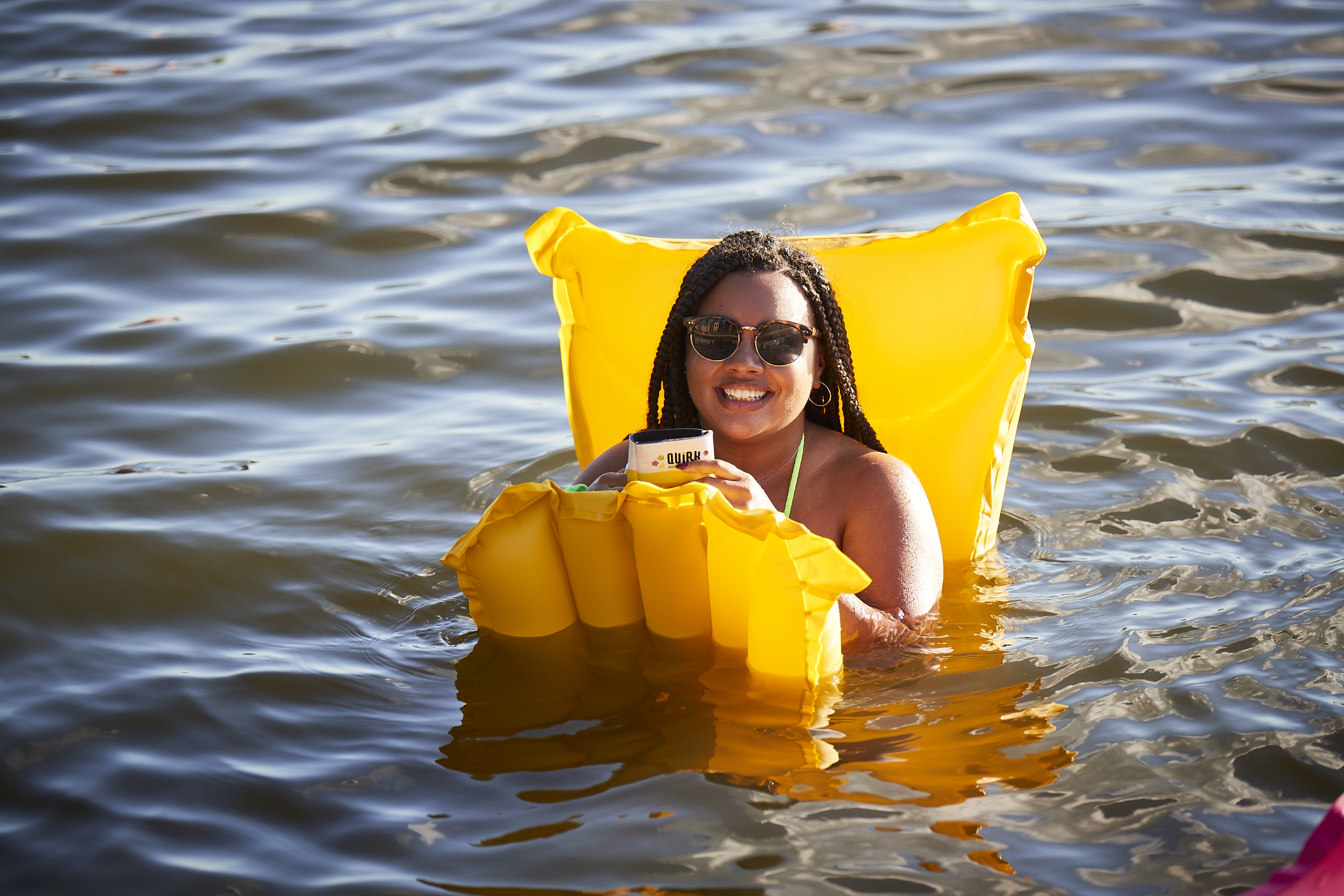 A woman with braided hair and sunglasses smiles while relaxing on a yellow Boolevard inflatable pool float in the water, holding a drink. The sunlight reflects off the water around her.