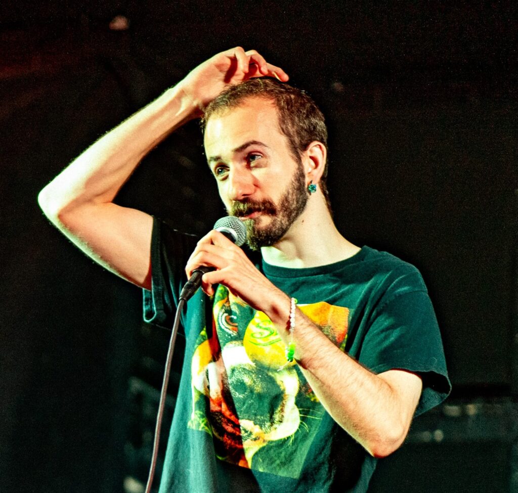 A man with a beard holds a microphone and scratches his head while speaking on stage. He wears a black T-shirt with a colorful graphic and has a bracelet on his wrist. The background is dark and out of focus.