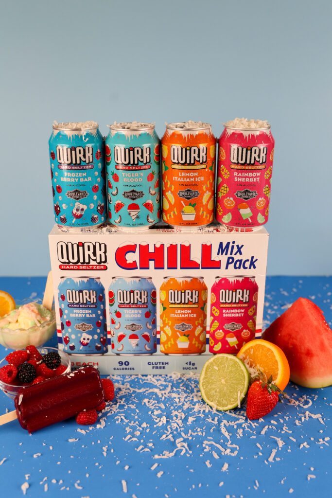 Five colorful cans of Quirk Hard Seltzer are displayed on top of a Chill Mix Pack box, surrounded by fruit slices, popsicles, berries, grated coconut, and a scoop of ice cream, against a blue background.