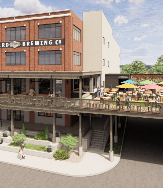 A brick building with a sign that reads "Boulevard Brewing Co" and “Brewhouse One,” featuring a large outdoor patio with colorful umbrellas, tables, and people; trees and parked cars are visible in the background.