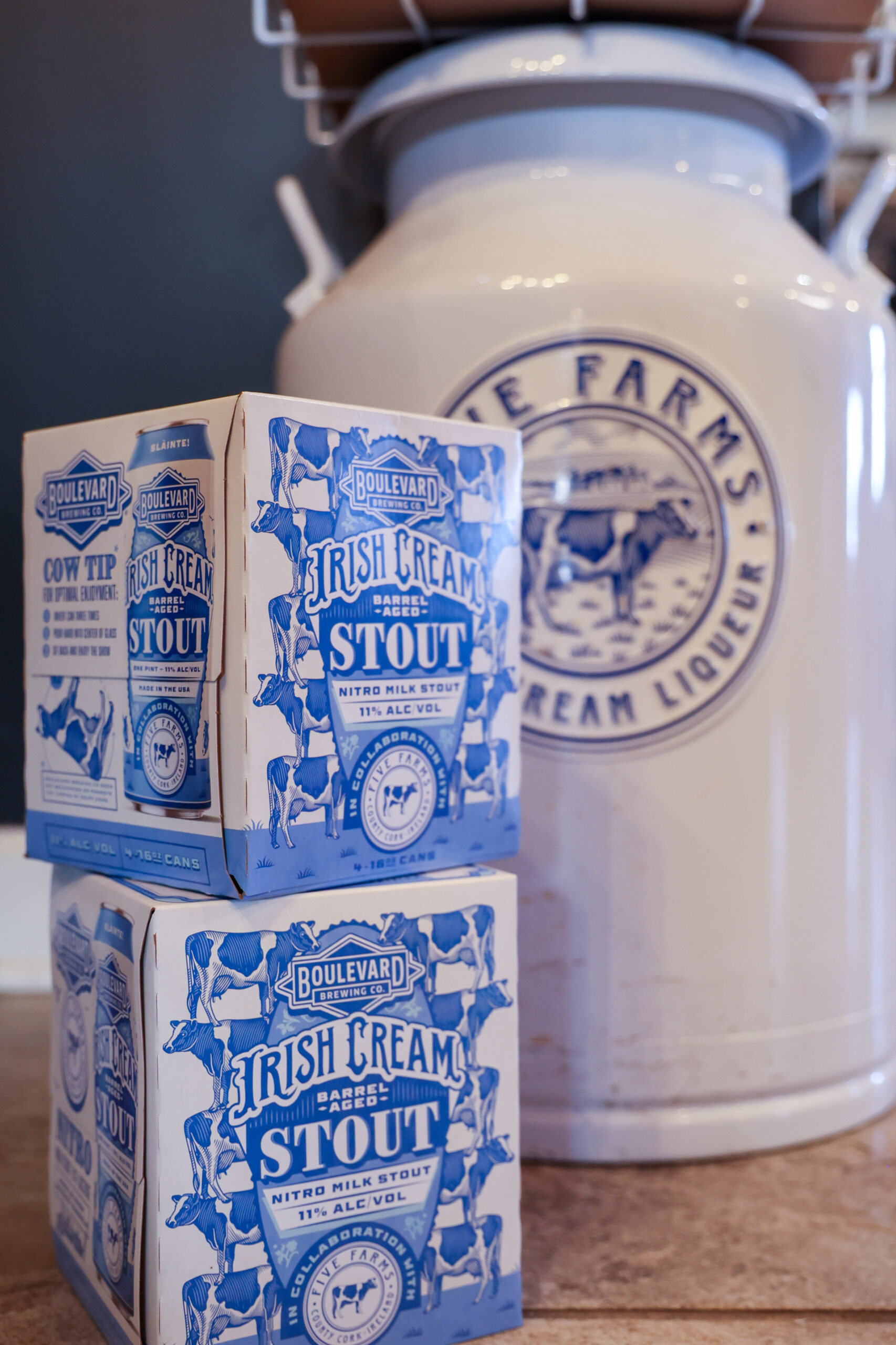 Two four-packs of Boulevard Brewing Co. Irish Cream Stout with blue and white packaging are stacked in front of a large vintage-style white milk can labeled "Farms Dairy Cream Liquor.