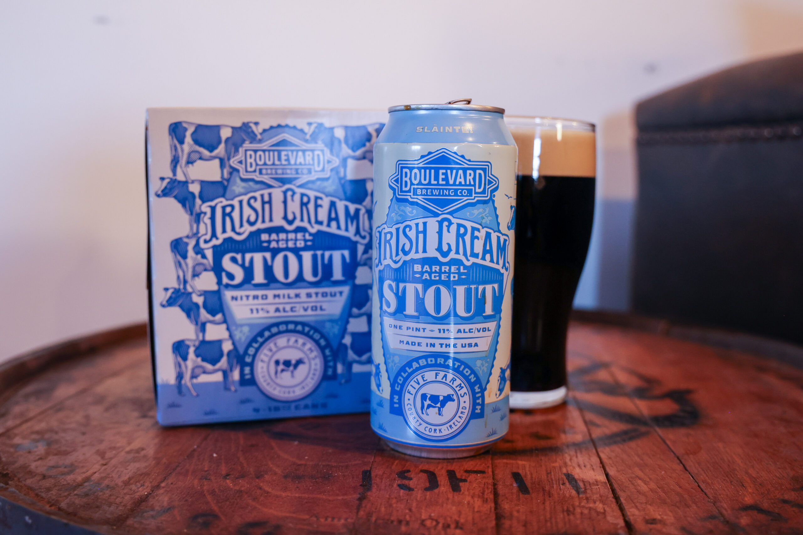A can and box of Boulevard Brewing Co. Irish Cream Barrel-Aged Stout sit on a wooden barrel next to a glass of dark stout beer with a creamy head. The packaging is blue and white with bold lettering.