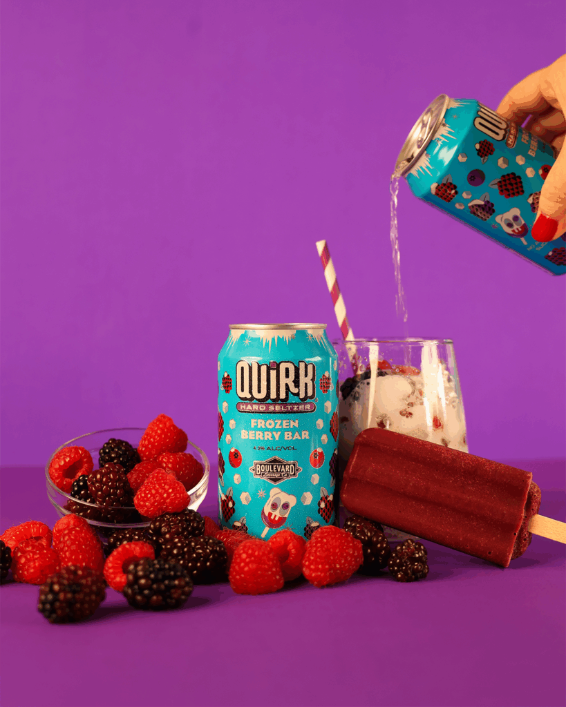 A can of Quirk Hard Seltzer sits among bowls of fresh blackberries and raspberries, a berry popsicle, and a glass with ice as another can pours seltzer into it, all set against a purple background.