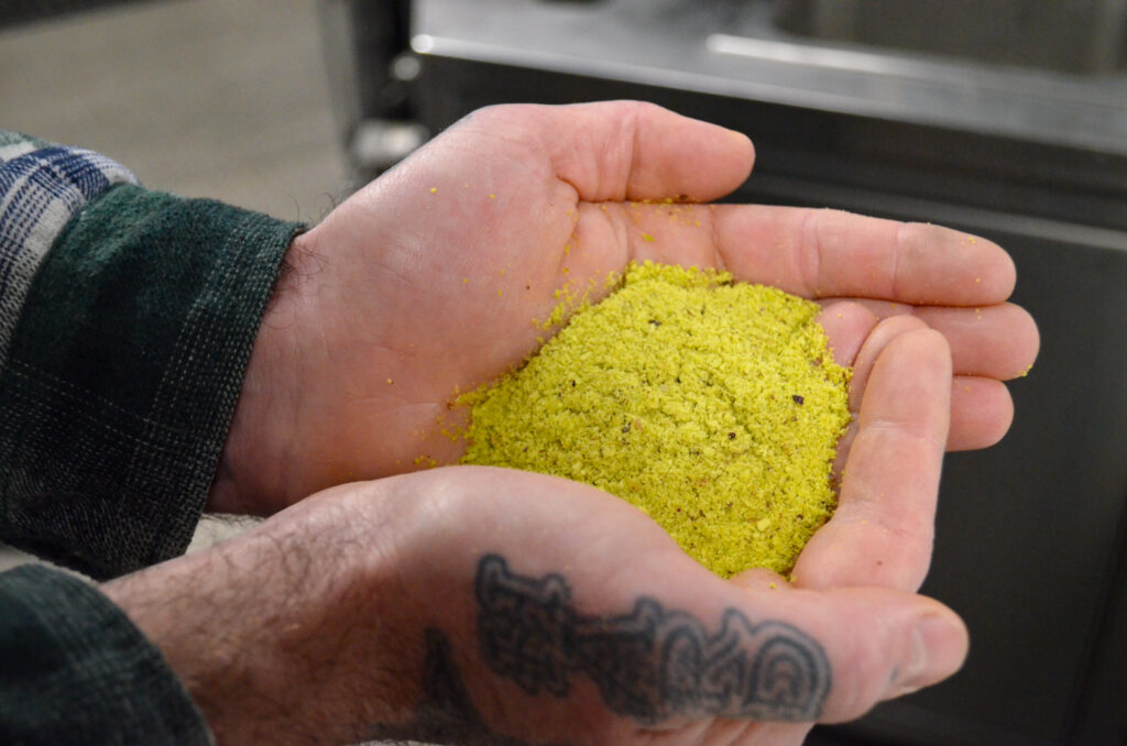 A person with a tattoo on their wrist holds a mound of yellow-green powder or spice in their cupped hands, evoking the artisanal spirit of brewhouse one, with a blurred kitchen background.