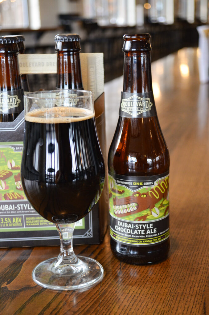 A bottle of Boulevard Brewing Co. Dubai-Style Chocolate Ale from their Brewhouse One series sits next to a filled glass on a wooden table, with a six-pack carrier in the background.