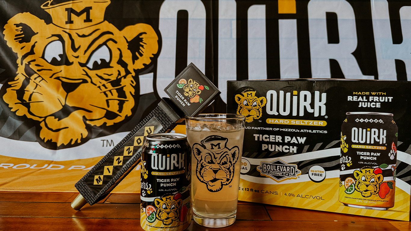 A glass of Tiger Paw Punch Quirk Hard Seltzer, a can, a 12-pack box, and Mizzou-themed items are displayed on a table in front of a large Missouri Tigers logo backdrop.