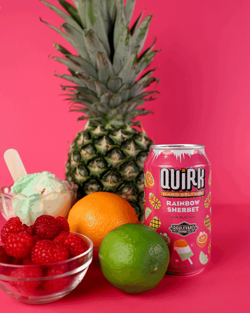 A can of Quirk Rainbow Sherbet hard seltzer stands on a pink surface, surrounded by a pineapple, orange, lime, bowl of raspberries, and bowl of ice cream with a spoon. The background is also bright pink.