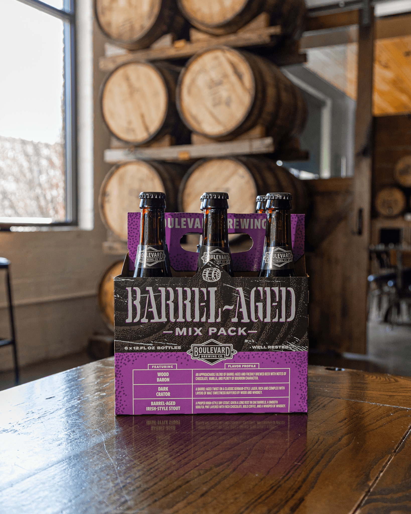 A four-pack of Boulevard Brewing Barrel-Aged Mix Pack beer bottles sits on a wooden table in a brewery, with large wooden barrels stacked in the background.
