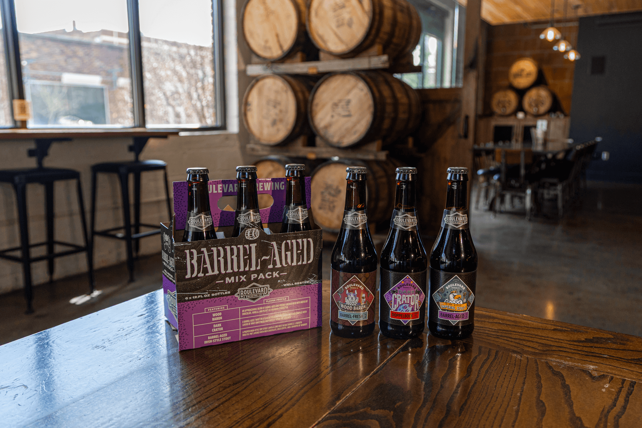 Four craft beer bottles and a purple six-pack carrier labeled "Barrel-Aged Mix Pack" sit on a wooden table in a brewery, with barrels and seating visible in the background.