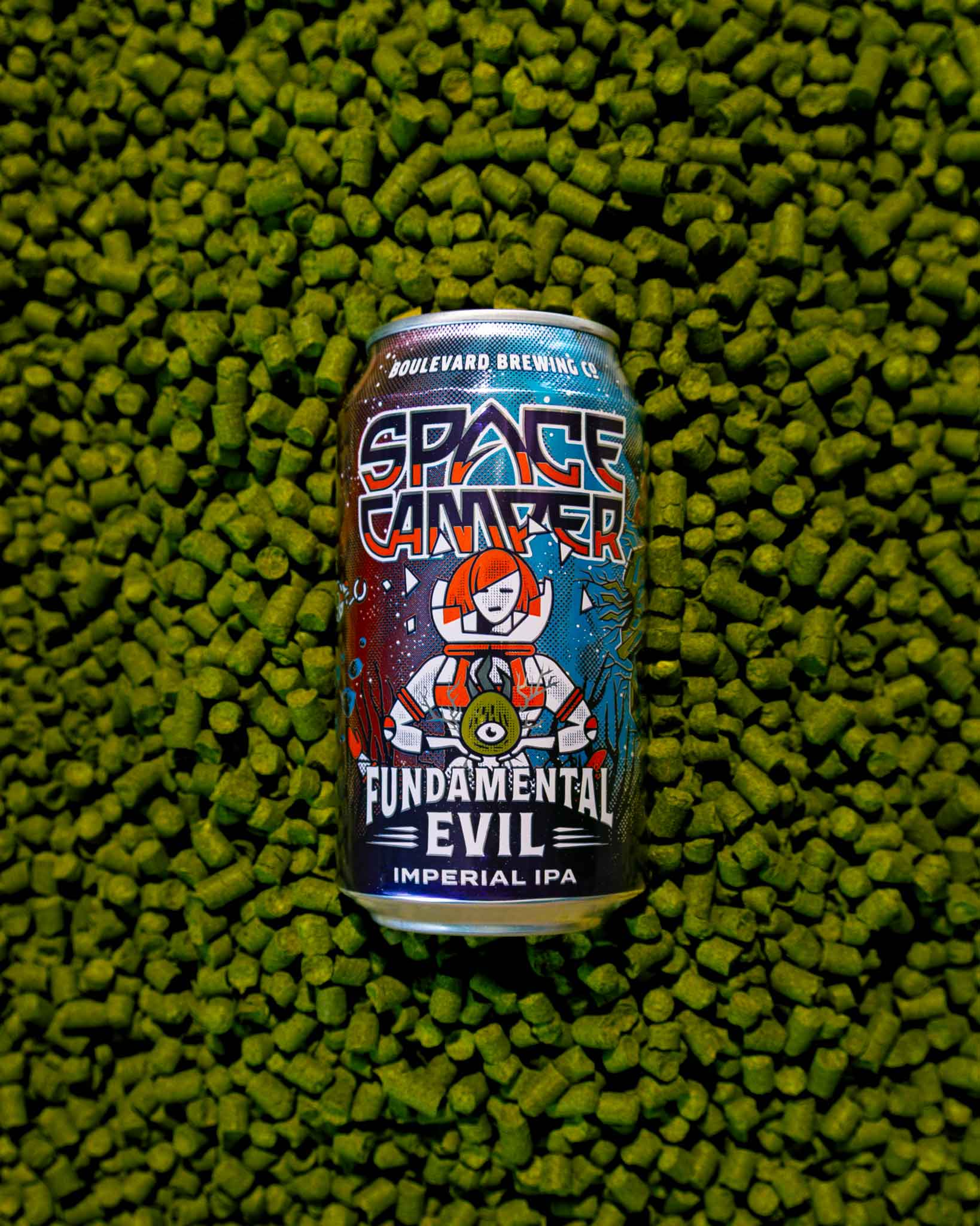 A can of Boulevard Brewing Co. Space Camper Fundamental Evil Imperial IPA is placed on a surface covered with green hop pellets. The can features a colorful space-themed illustration.