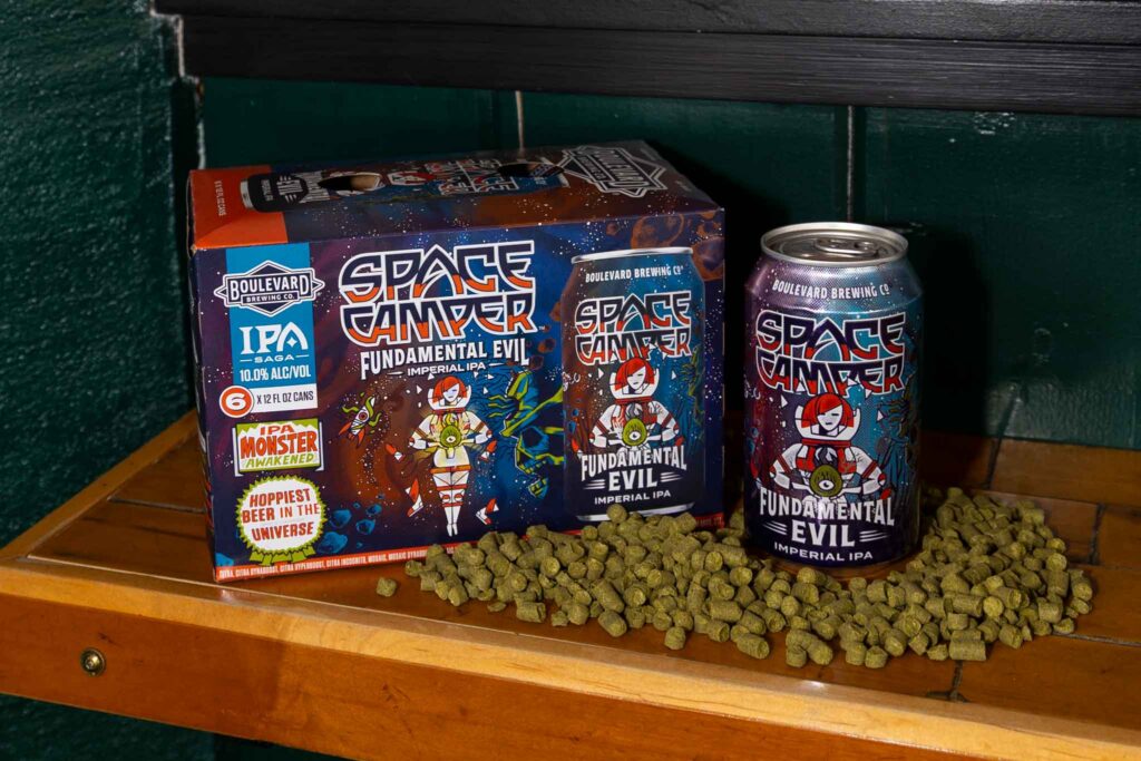 A 6-pack box and can of Space Camper Fundamental Evil Imperial IPA sit on a wooden shelf, surrounded by loose green hop pellets. The packaging features a cartoon astronaut cat and colorful space-themed artwork.