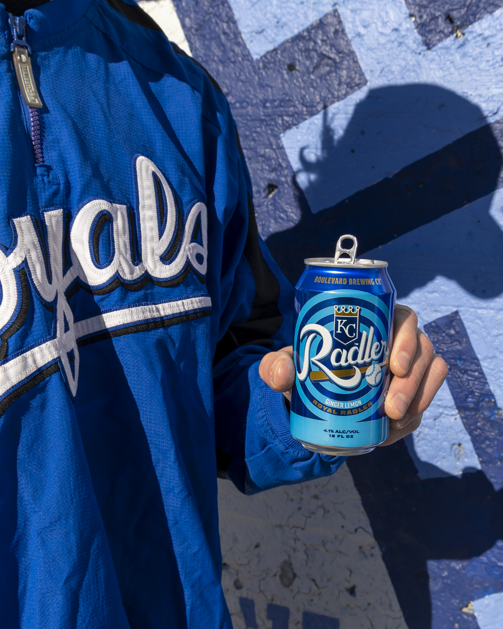 A person wearing a blue Royals jacket holds a can of Boulevard Brewing Co. Royal Radler against a blue and white painted wall in bright sunlight.