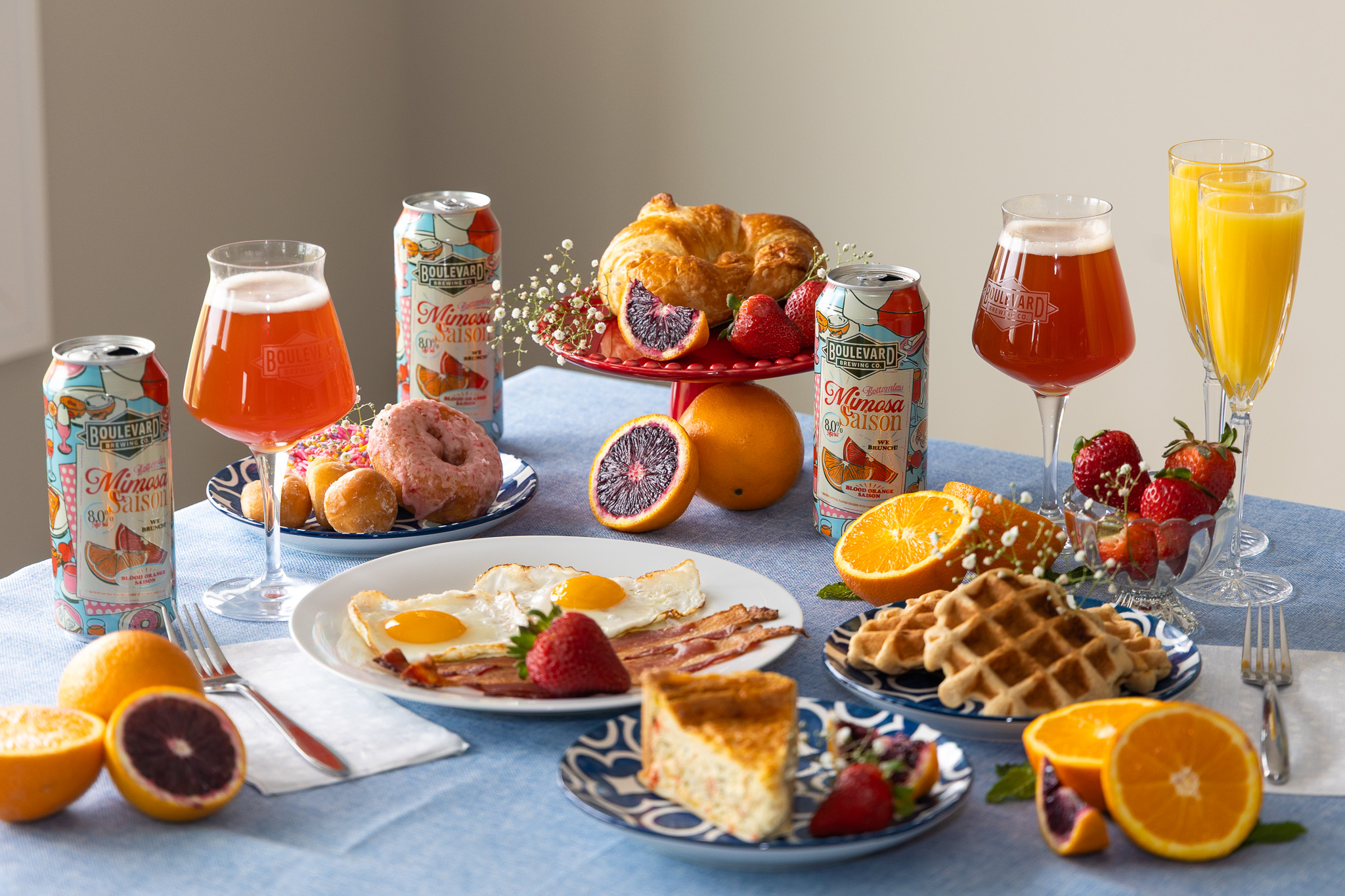 A brunch table with mimosas, cans of Mimosa Saison beer, croissants, waffles, eggs with bacon, quiche, donuts, strawberries, and sliced oranges on a blue tablecloth.