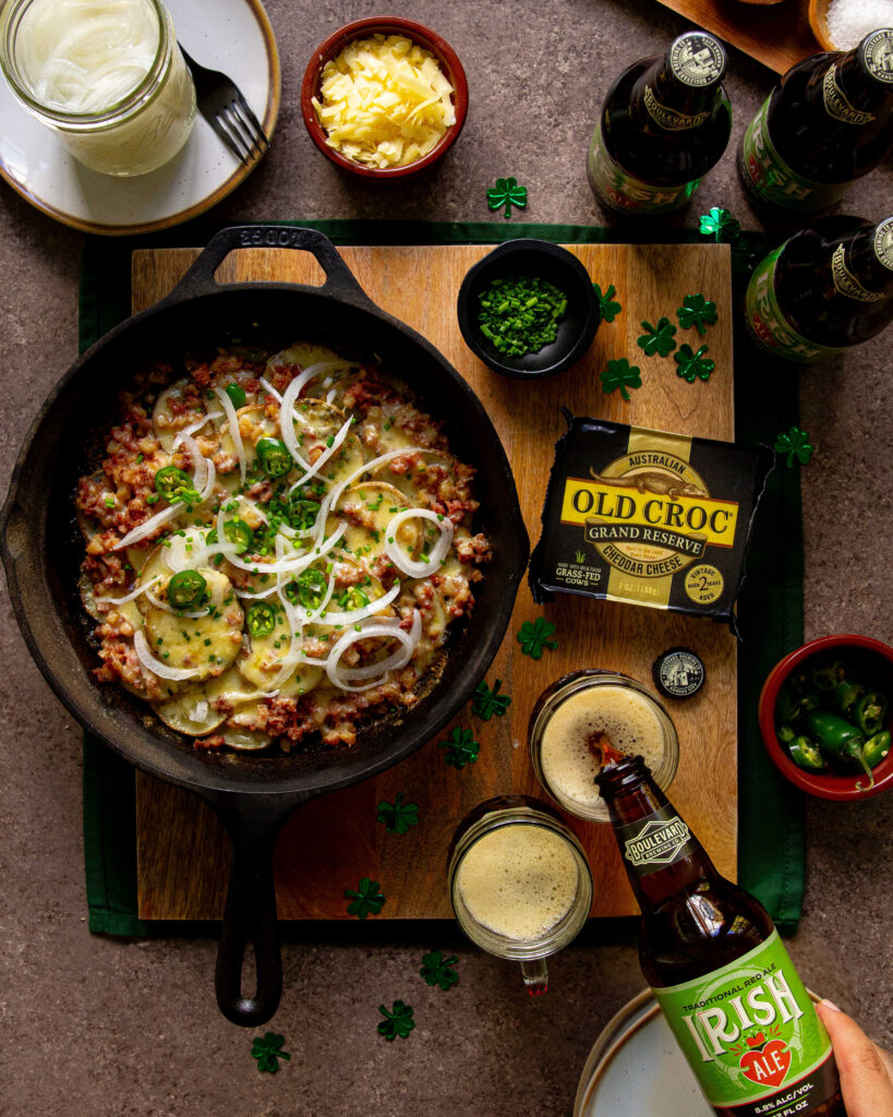 A skillet of cheesy, onion-topped ground beef sits on a wooden board with a box of Old Croc cheese, chopped herbs, shredded cheese, beer bottles, and a hand pouring Irish ale, surrounded by green shamrock confetti.