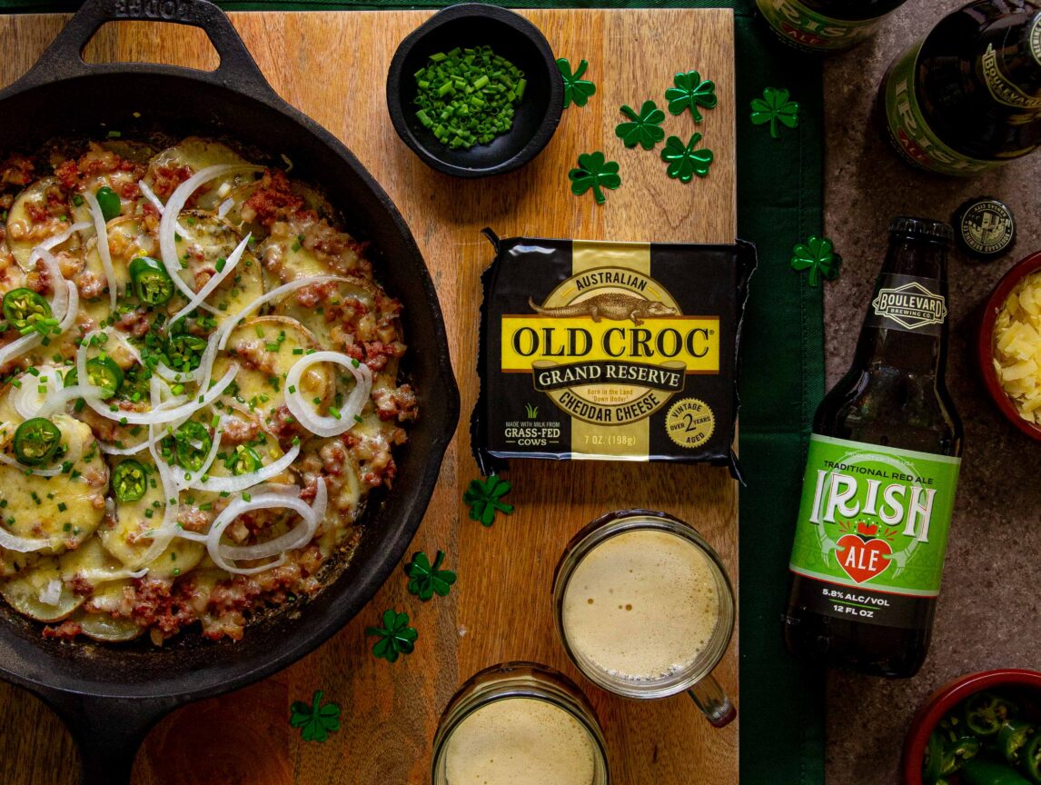 A skillet with a cheesy potato and meat dish is surrounded by bottles of Irish ale, a block of Old Croc cheese, two mugs of beer, small bowls of chives and sauces, and paper shamrocks on a wooden board.