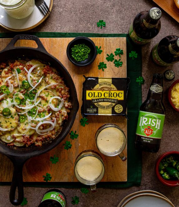 A skillet with a cheesy potato and meat dish is surrounded by bottles of Irish ale, a block of Old Croc cheese, two mugs of beer, small bowls of chives and sauces, and paper shamrocks on a wooden board.