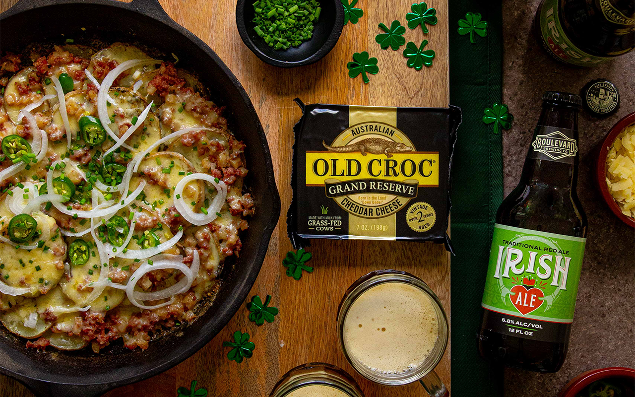 A skillet of cheesy potato dish topped with sliced onions and jalapeños sits beside a block of Old Croc cheddar, chopped chives, shredded cheese, shamrock decor, and a glass and bottle of Irish ale—perfect for any Boulevard Brewing Irish Ale recipe.