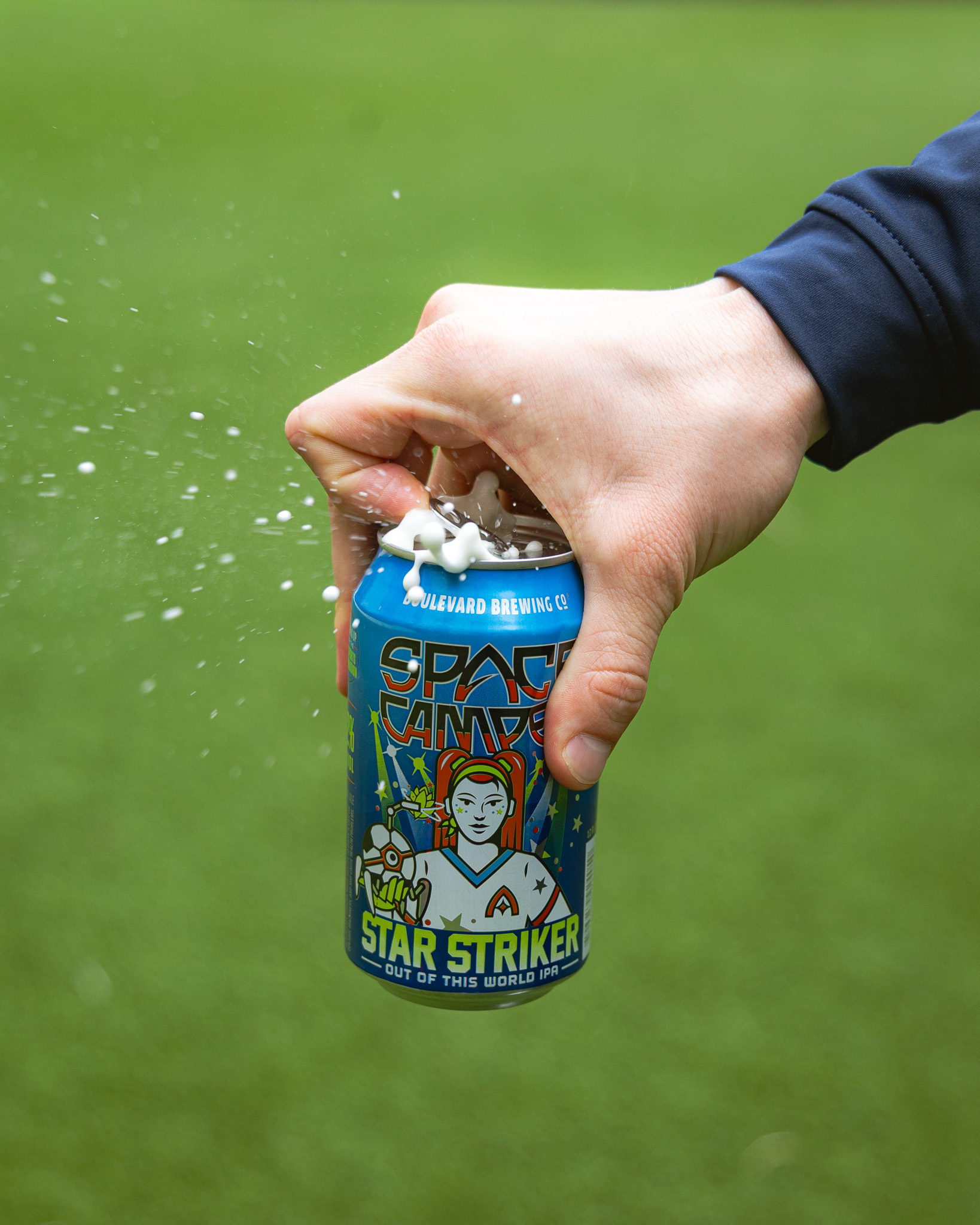 A hand is opening a can of Space Camper Star Striker beer, causing foam to spray out, with a grassy background. The can is blue and features colorful artwork of a person in a spacesuit.