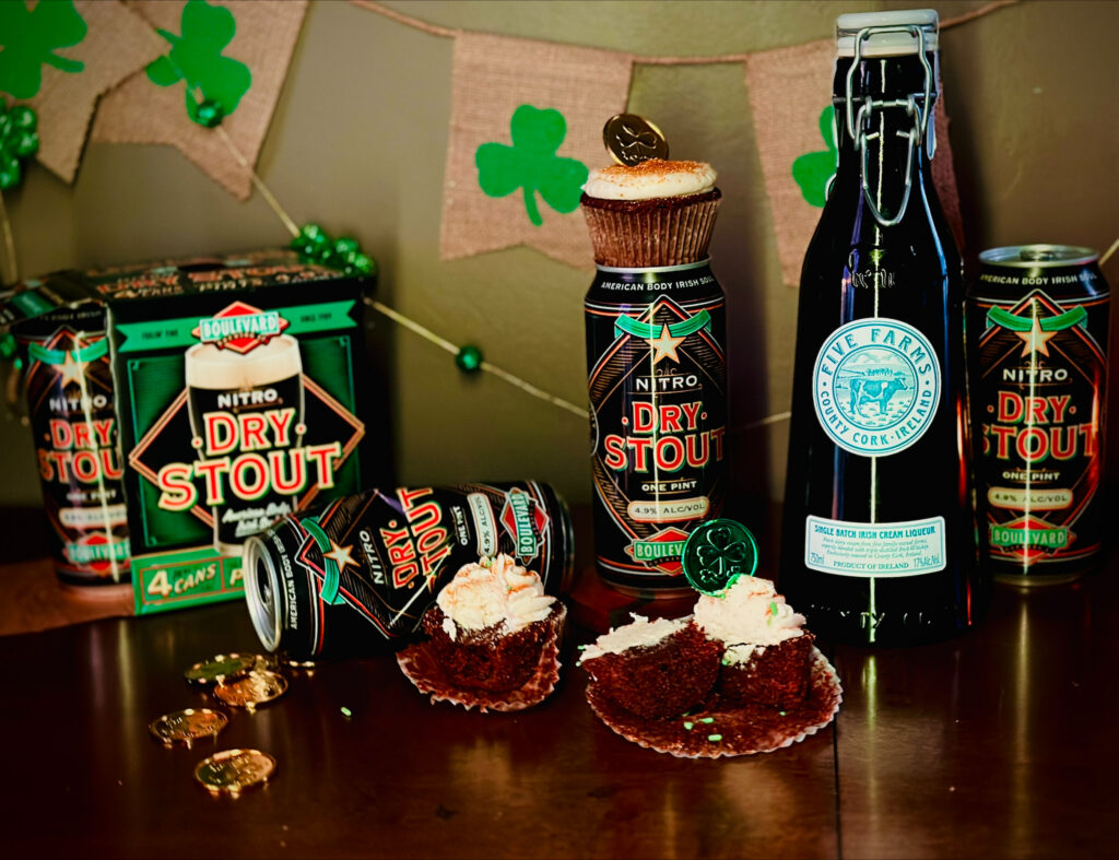 A St. Patrick’s Day-themed display features Dry Stout beer cans, a bottle of Irish cream liqueur, chocolate cupcakes with cream frosting, gold coins, shamrock decorations, and a Boulevard Brewing Irish Ale recipe on a wooden surface.
