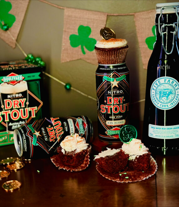 A St. Patrick’s Day-themed display features Dry Stout beer cans, a bottle of Irish cream liqueur, chocolate cupcakes with cream frosting, gold coins, shamrock decorations, and a Boulevard Brewing Irish Ale recipe on a wooden surface.