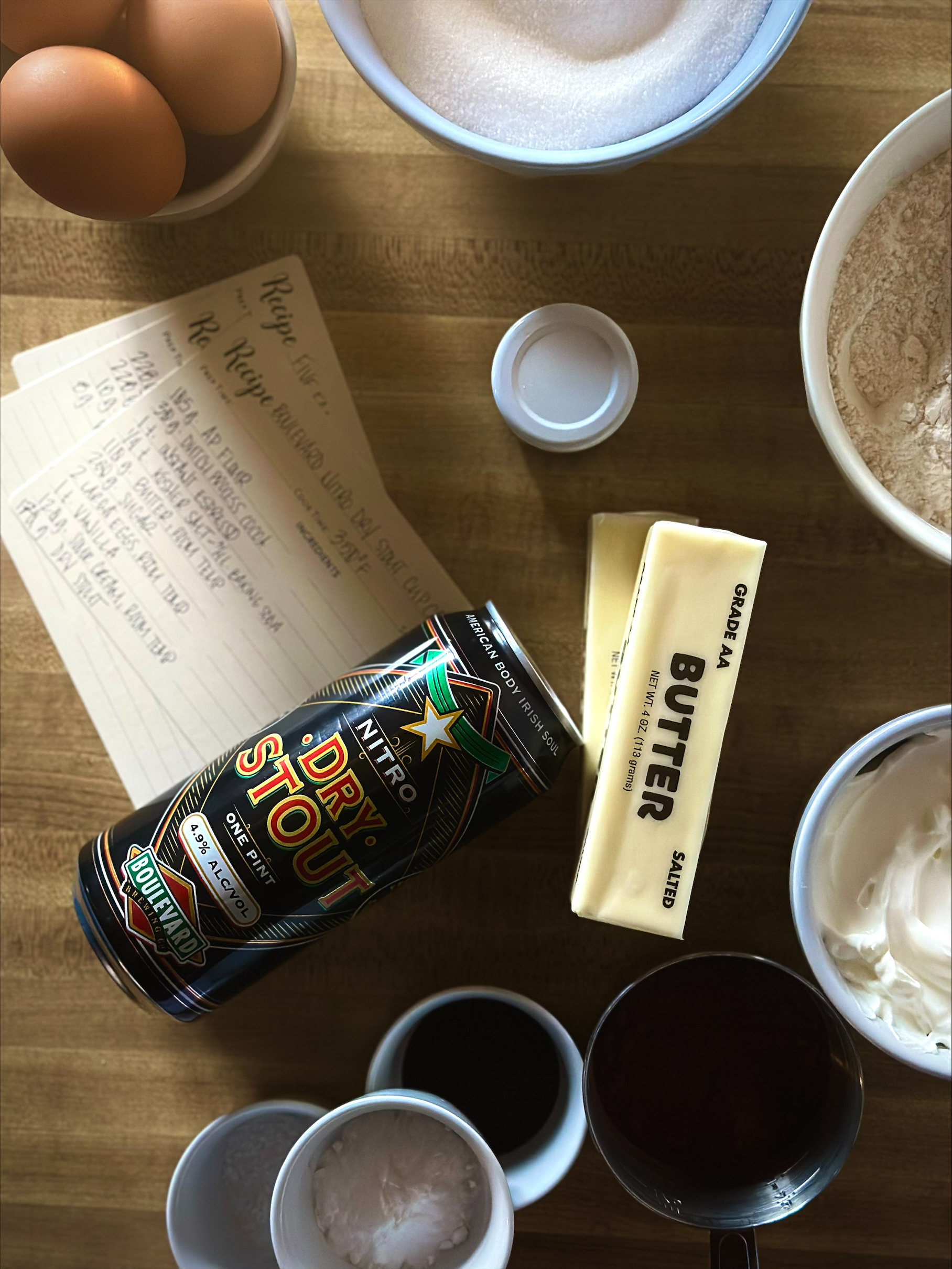 Baking ingredients on a wooden counter, including eggs, sugar, flour, butter sticks, sour cream, vanilla, cocoa powder, and a can of dry stout—plus handwritten recipe cards featuring a Boulevard Brewing Irish Ale recipe.