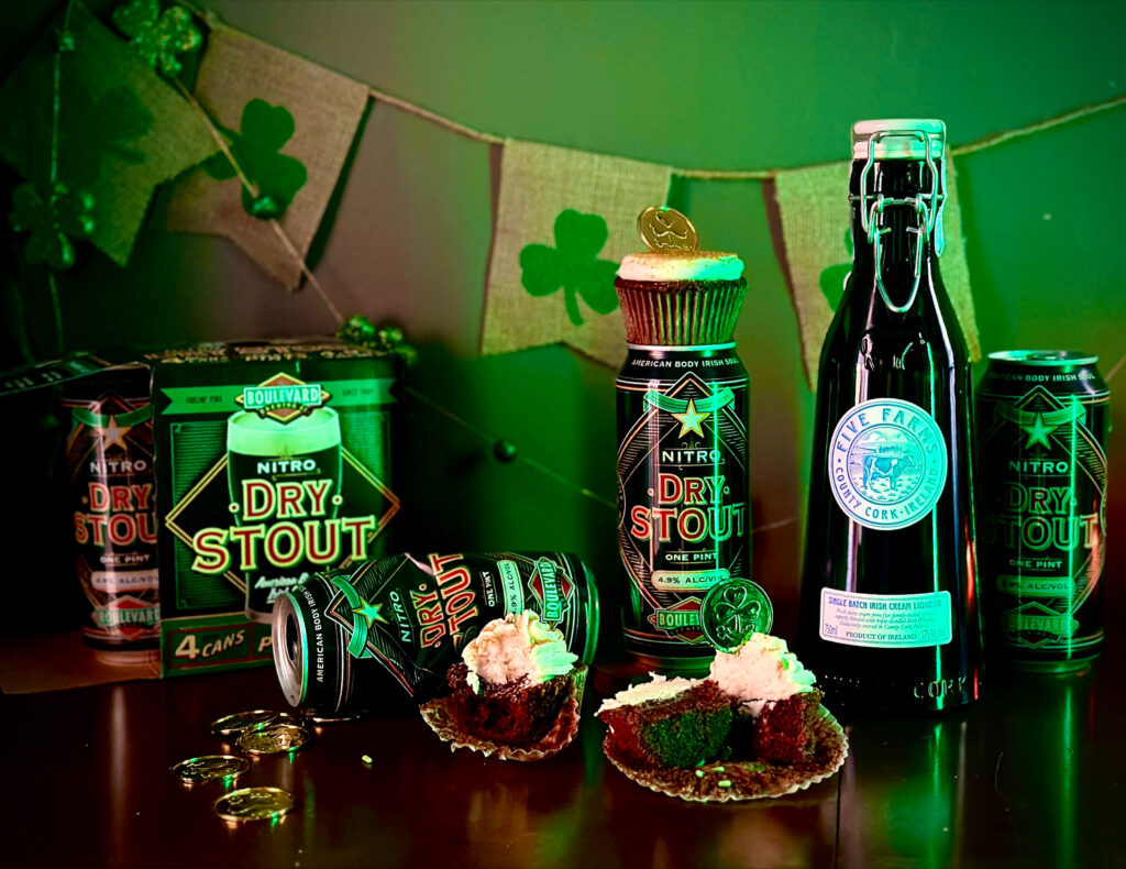 A St. Patrick’s Day-themed setup features Dry Stout beer cans, a bottle of stout, gold chocolate coins, cupcakes with whipped cream, and a shamrock garland under green lighting—perfect inspiration for your own Boulevard Brewing Irish Ale recipe celebration.