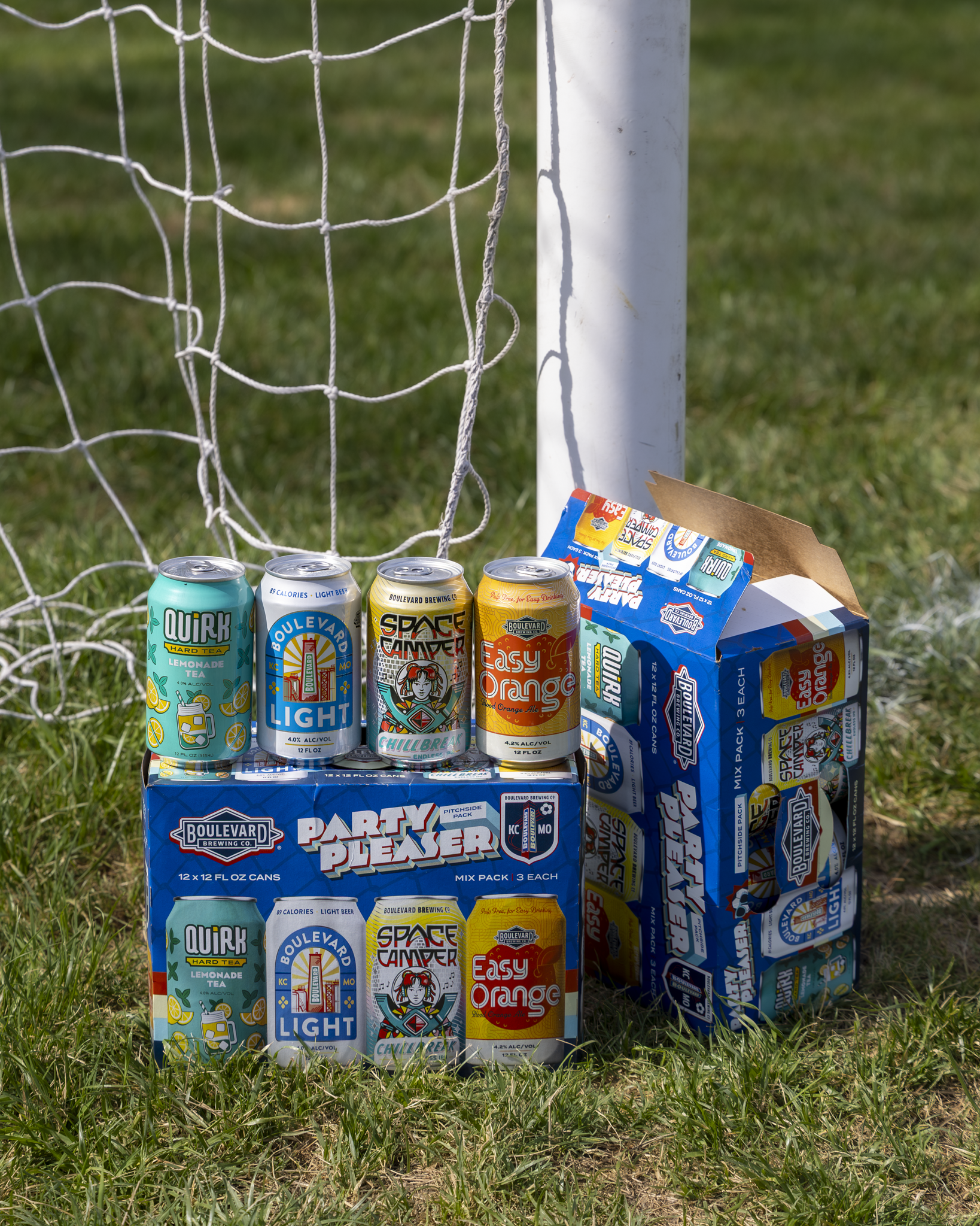Four colorful cans of Boulevard Brewing Co. Kansas City beer and seltzer are stacked by a soccer net on grass, next to an open 12-pack box featuring matching can designs—a lively scene from Missouri's hometown brewery.