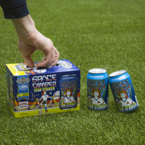 A hand reaches for a box of Space Camper Star Striker IPA on green grass, with two cans of the colorful, space-themed beer placed next to the box.