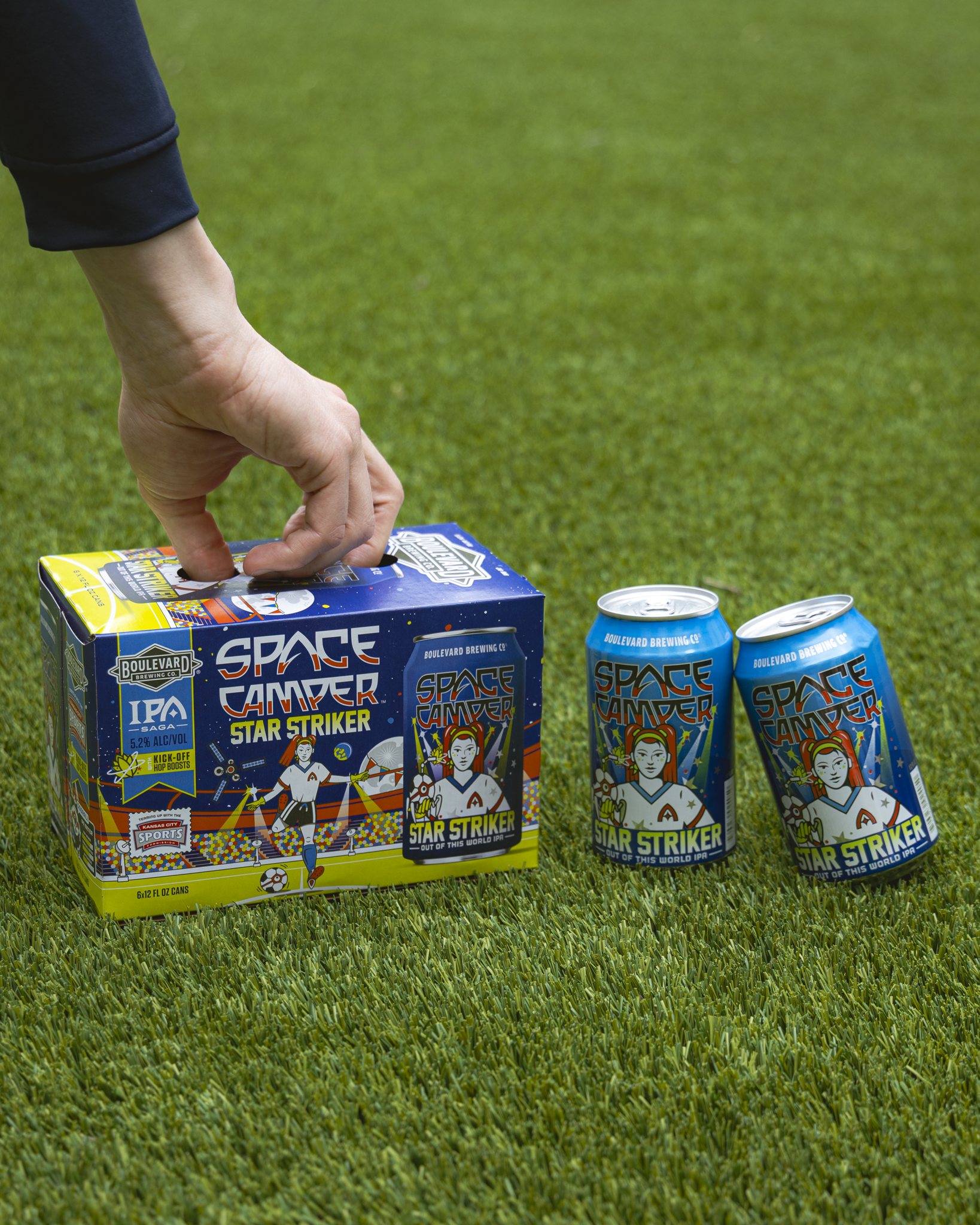A hand reaches for a box of Space Camper Star Striker IPA on green grass, with two cans of the colorful, space-themed beer placed next to the box.
