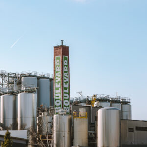 An industrial brewery complex with large silver fermentation tanks and a tall brick chimney that has "Boulevard" painted on it in green and white letters, set against a clear blue sky.