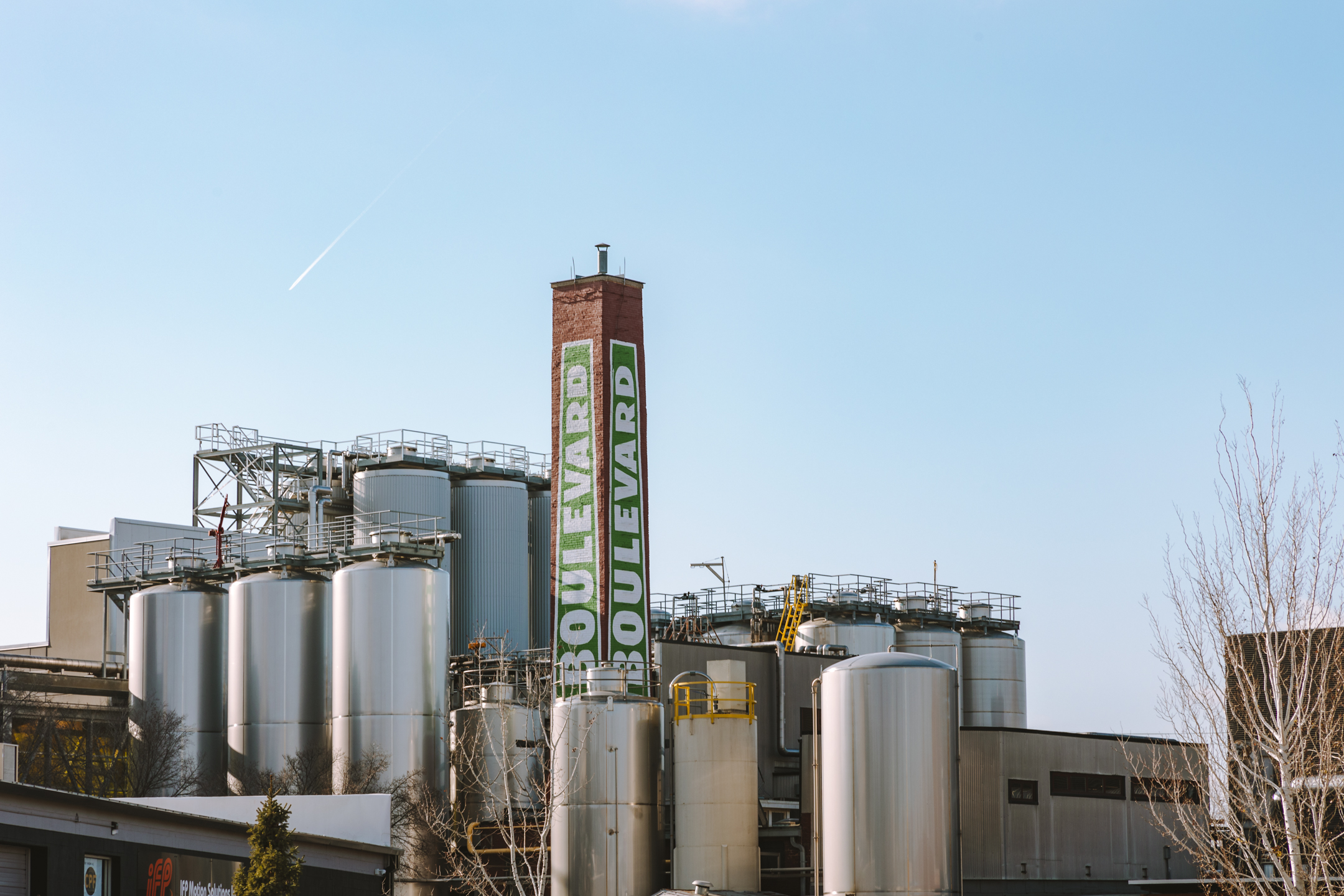 An industrial brewery complex with large silver fermentation tanks and a tall brick chimney that has "Boulevard" painted on it in green and white letters, set against a clear blue sky.