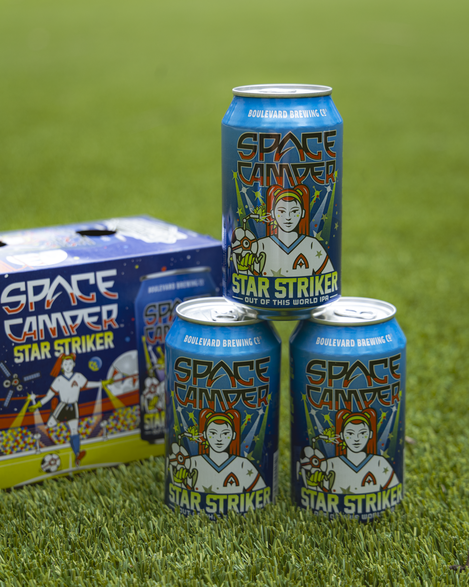 Three cans and a six-pack box of "Space Camper Star Striker" beer by Boulevard Brewing Co. sit on green grass. The cans feature colorful space and soccer-themed artwork with a character in a spacesuit.