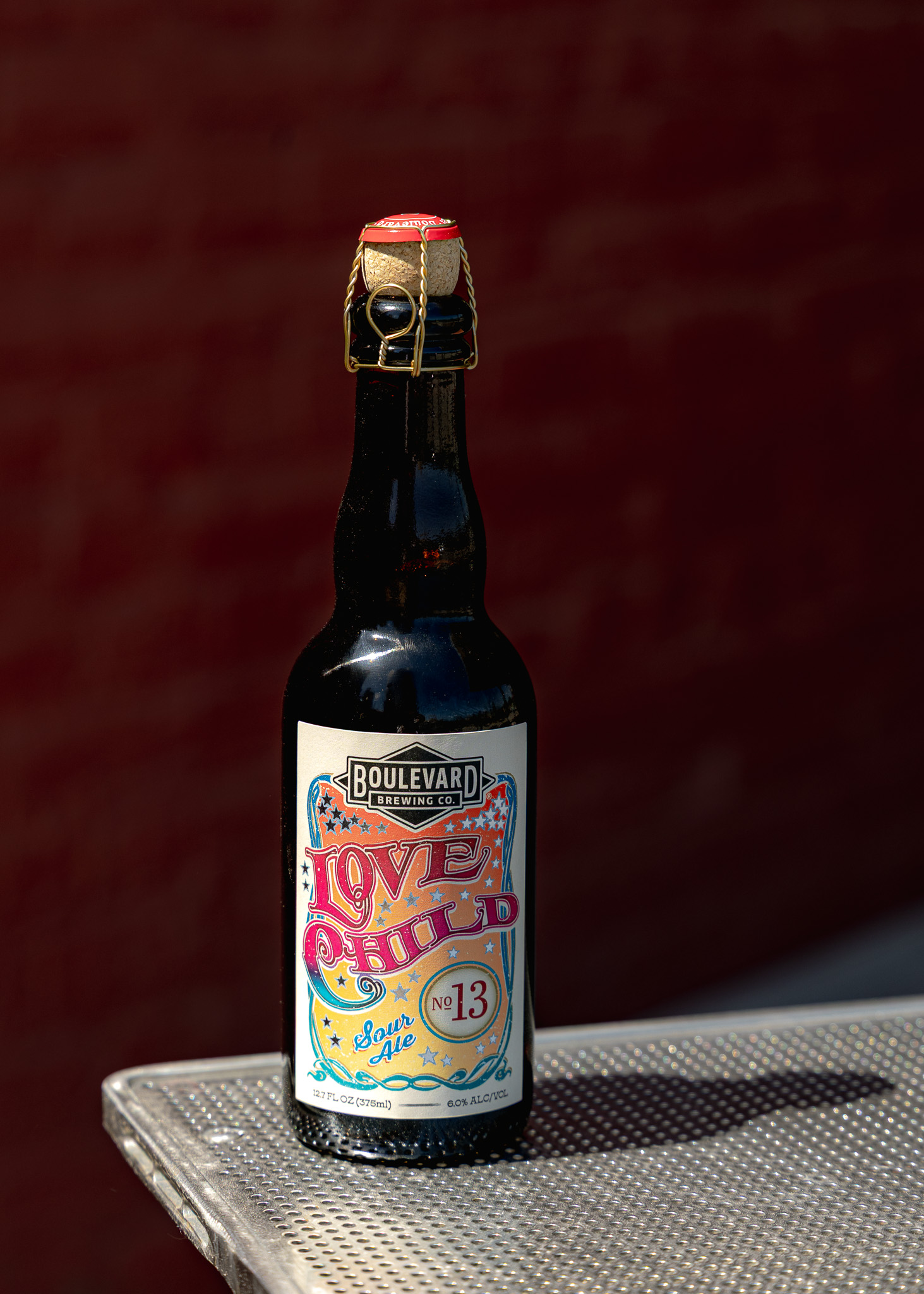 A dark bottle of Boulevard Brewing Co. Love Child No. 13 sour ale sits on a metal table, with a colorful label and a wired cork, against a blurred brick background.