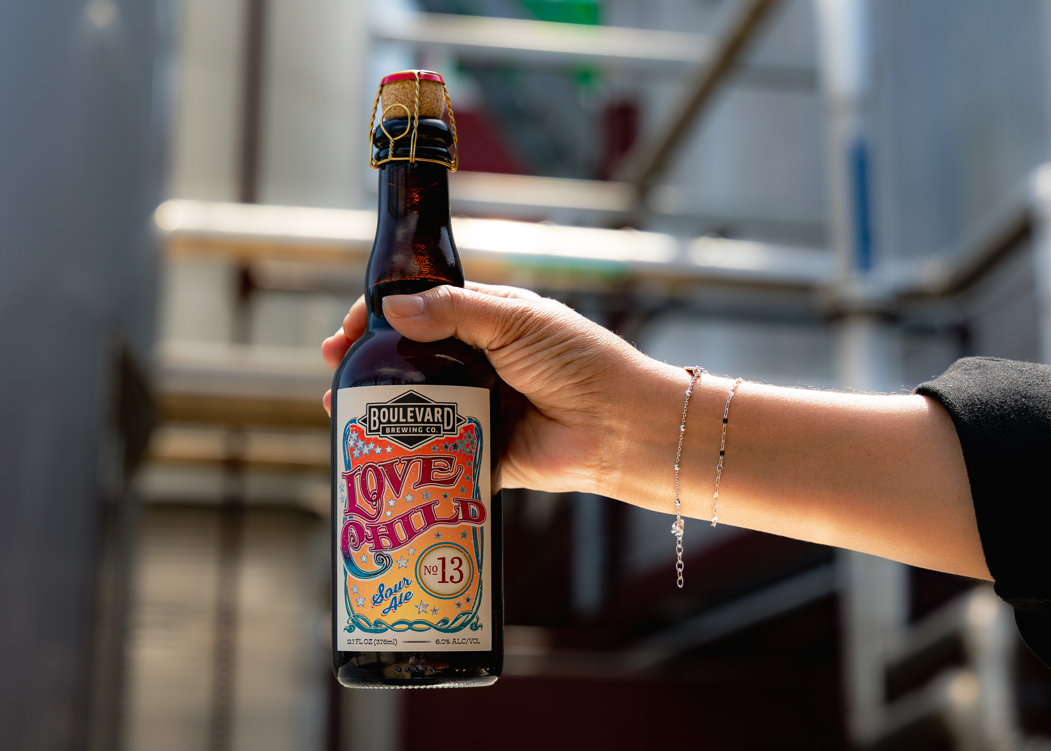 A person holding a bottle of Boulevard Brewing Co. Love Child No. 13 beer outdoors, with metal railings and blurred background. The person wears a delicate bracelet on their wrist.