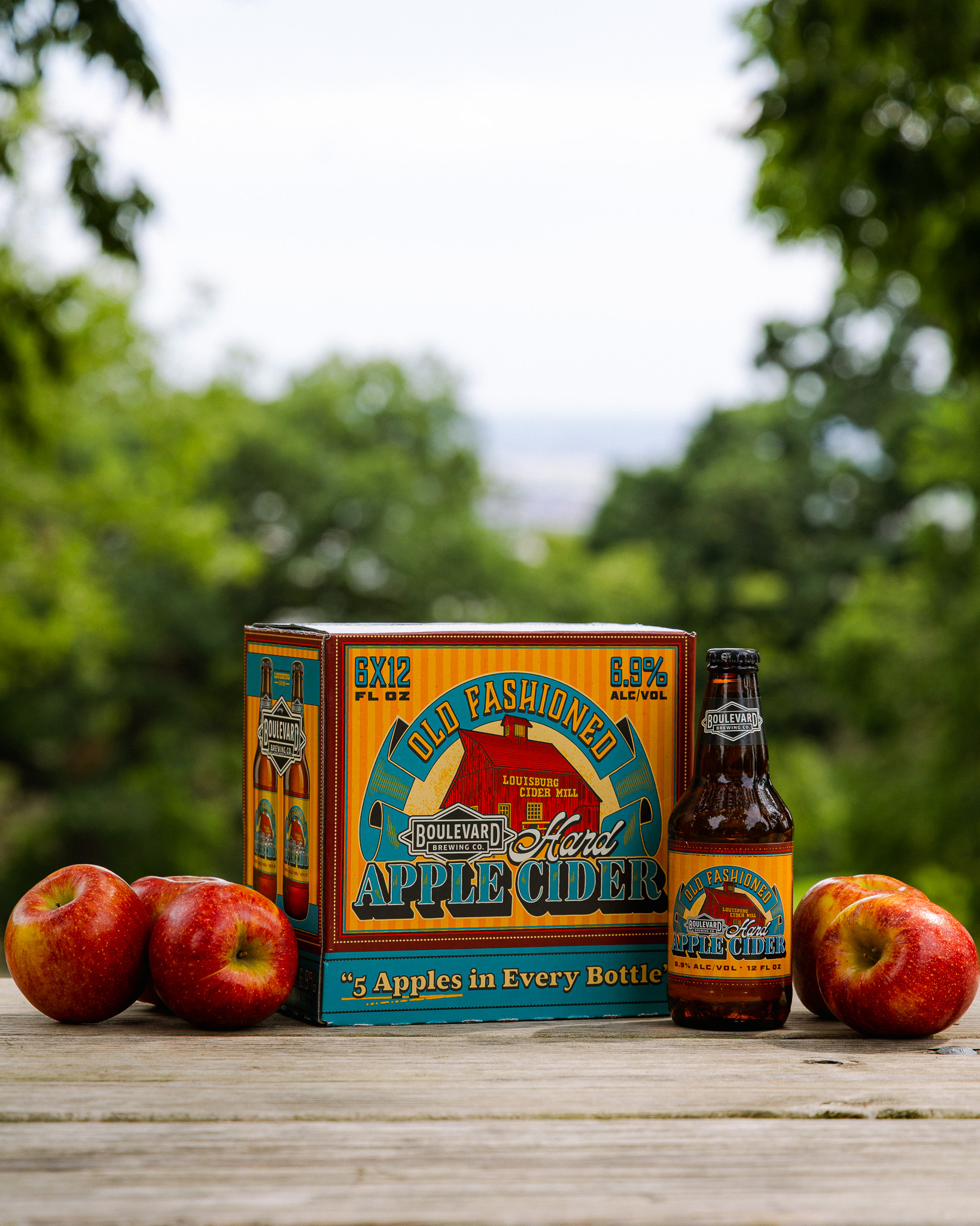 A box and bottle of Old Fashioned Hard Apple Cider are on a wooden table outdoors, flanked by red apples, with green trees and a blurry background visible.