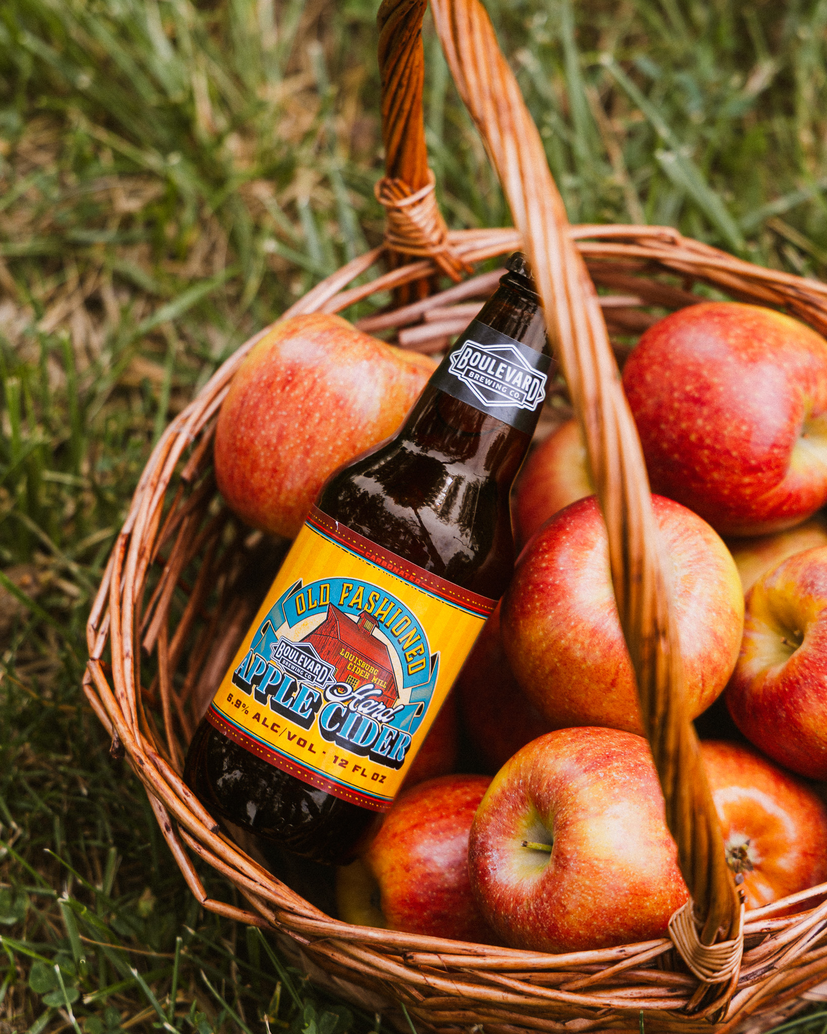 A bottle of Old Fashioned Hard Apple Cider rests in a basket filled with red apples on grass.