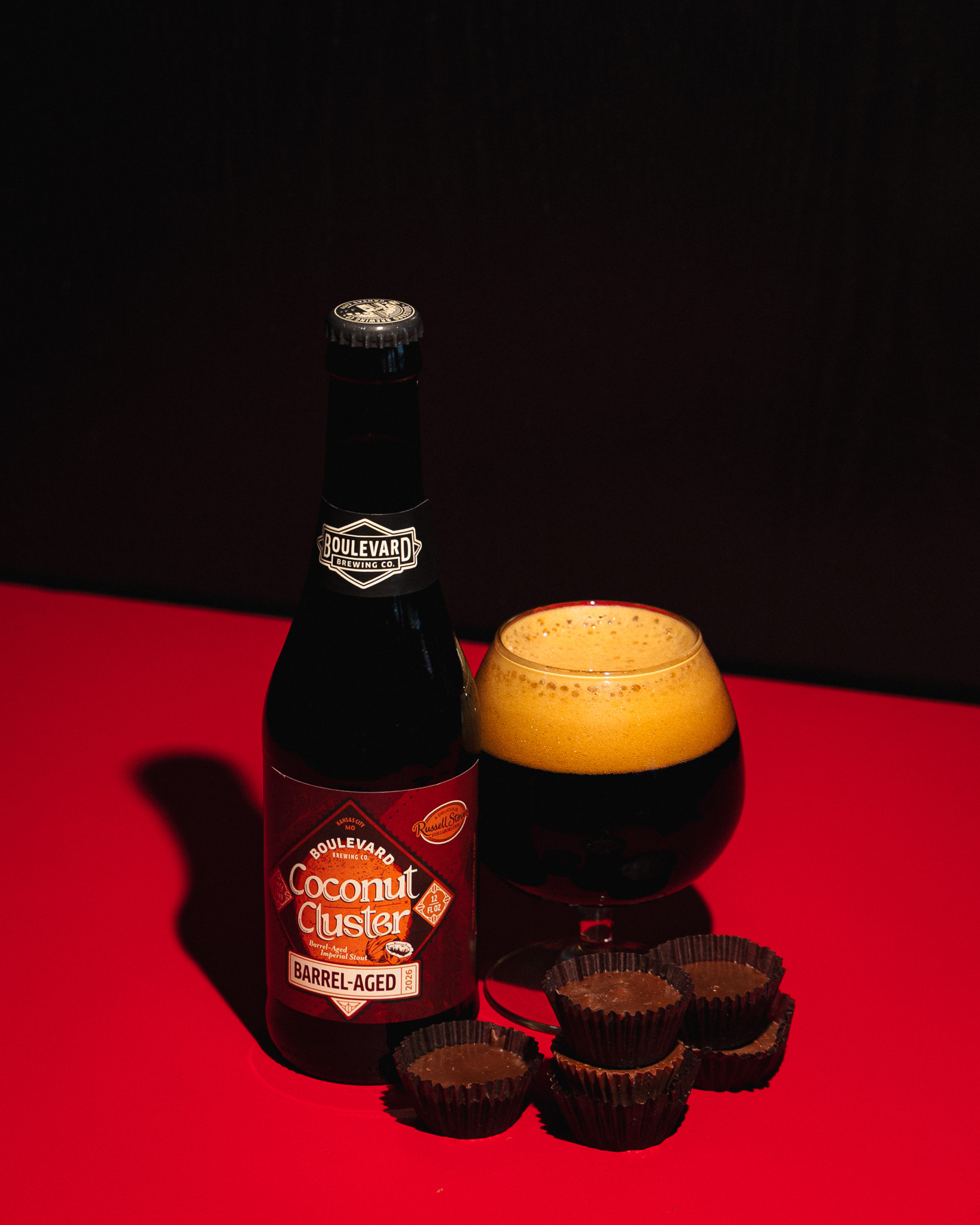 A bottle of Boulevard Coconut Custer Barrel-Aged beer, a glass filled with dark beer and foam, and several empty brown chocolate wrappers sit on a red surface against a dark background.