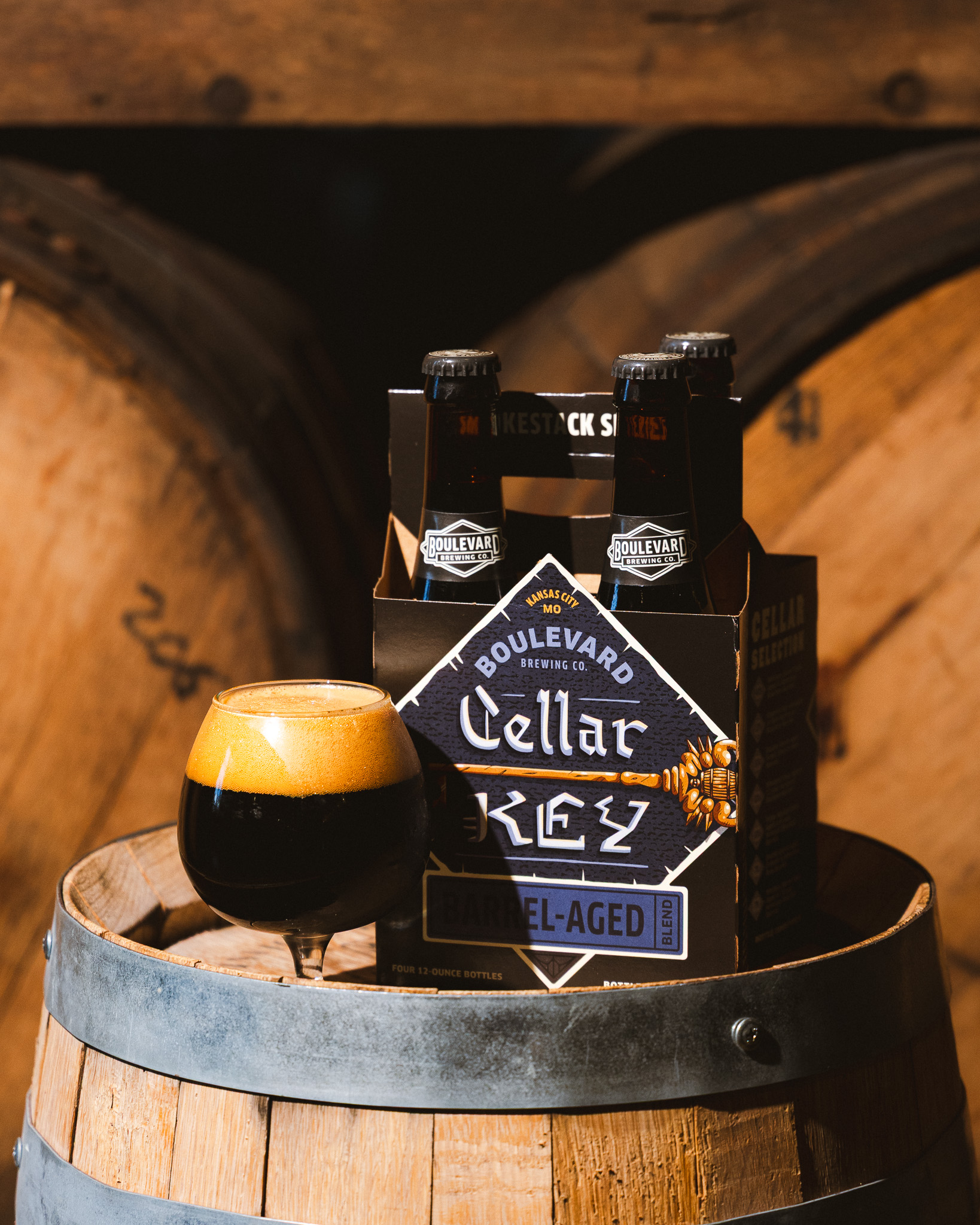 A pack of Boulevard Cellar Key barrel-aged beer and a glass of dark beer with foam sit on a wooden barrel, with more barrels blurred in the background.
