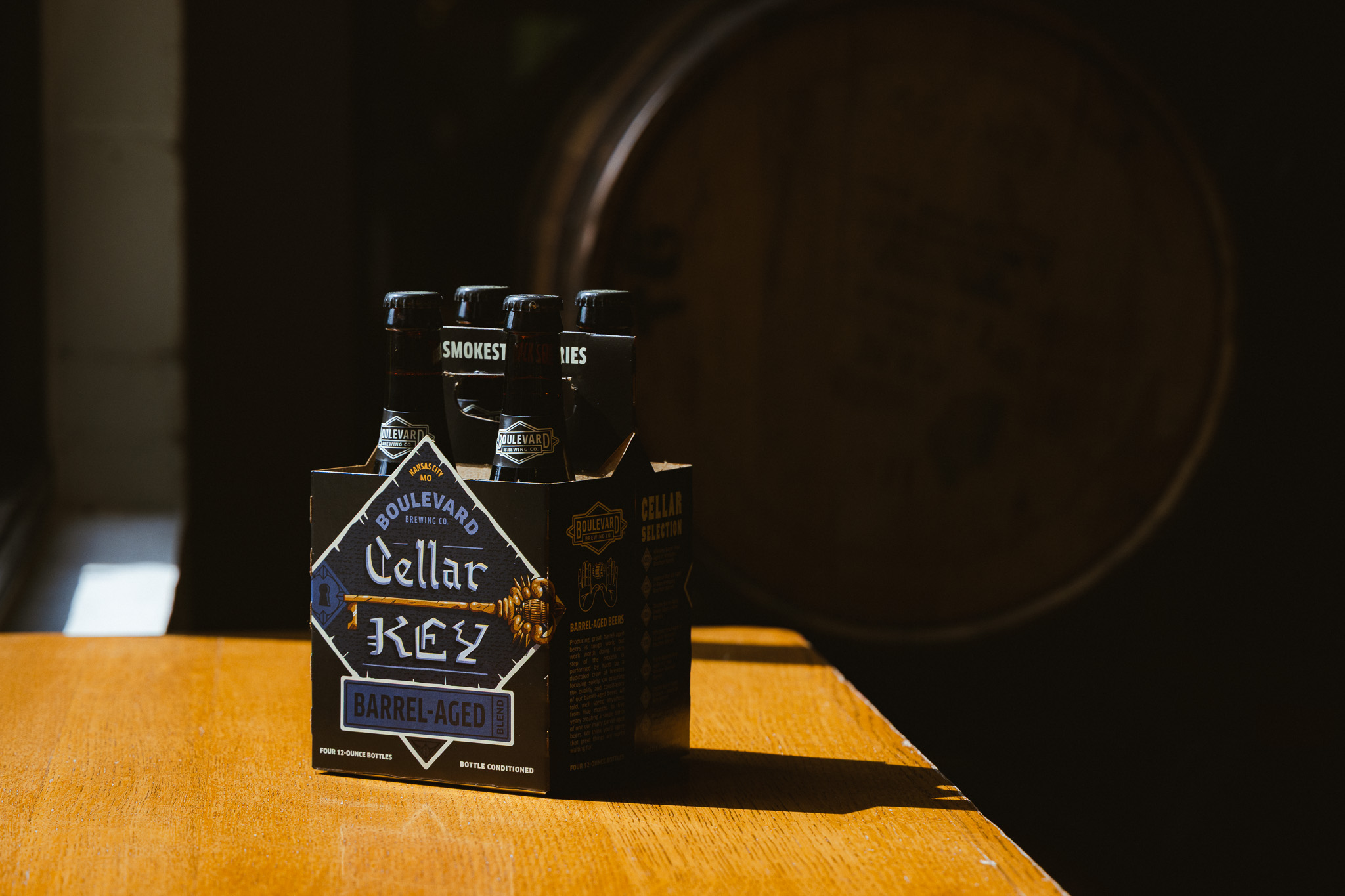 A four-pack of Boulevard Cellar Klez Barrel-Aged beer bottles sits on a wooden table in sunlight, with a barrel in the shadowy background.