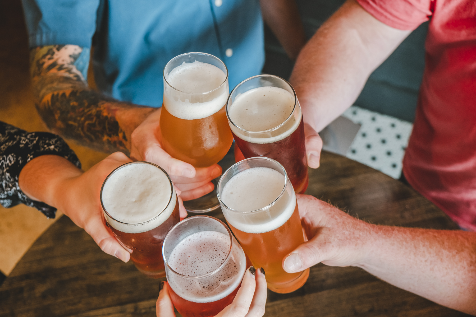Five people clinking glasses of beer together over a wooden table, seen from above. Four arms are visible, each holding a glass filled with frothy amber beer.